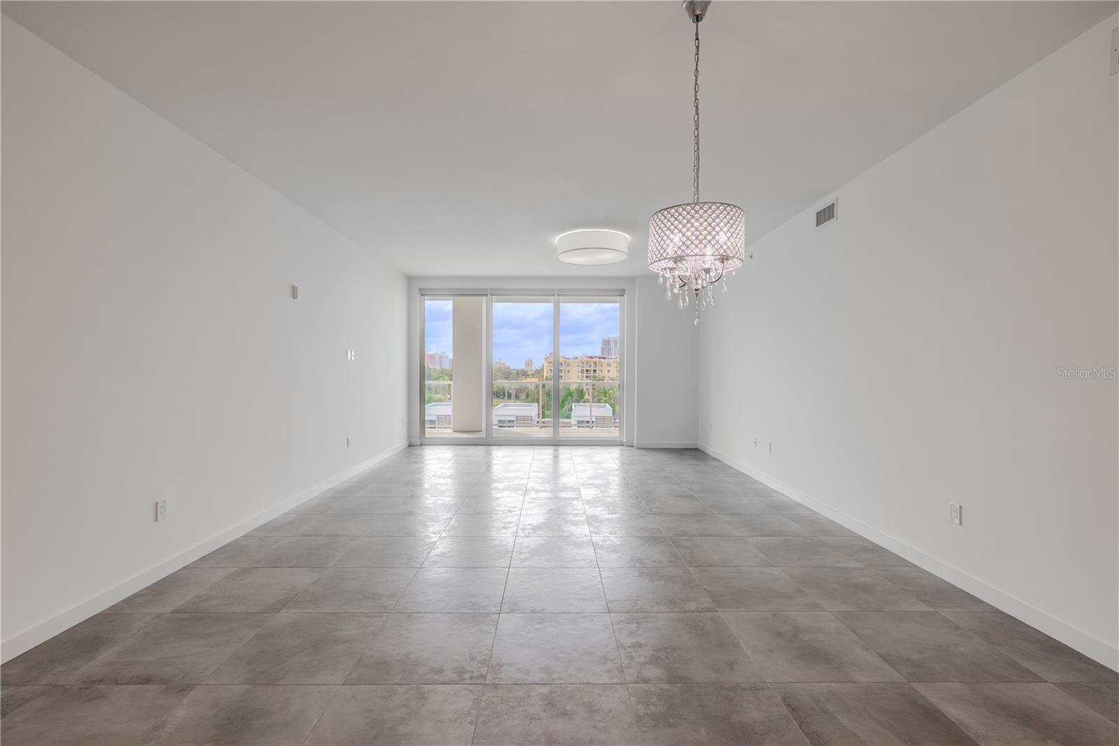 This bright and spacious dining and living area offers an elegant, open layout with sleek tile flooring and contemporary lighting. Floor-to-ceiling glass doors flood the space with natural light and open to a large balcony with sweeping water and skyline views. The modern chandelier adds a touch of sophistication, creating the perfect setting for entertaining or relaxing in style.