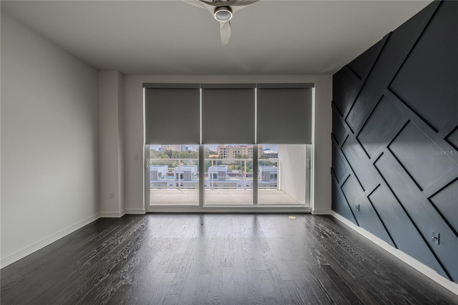 The serene primary bedroom offers a sophisticated retreat with direct balcony access and stunning water views. Rich hardwood floors and a custom geometric accent wall add warmth and modern flair, while floor-to-ceiling sliding glass doors fill the room with natural light.