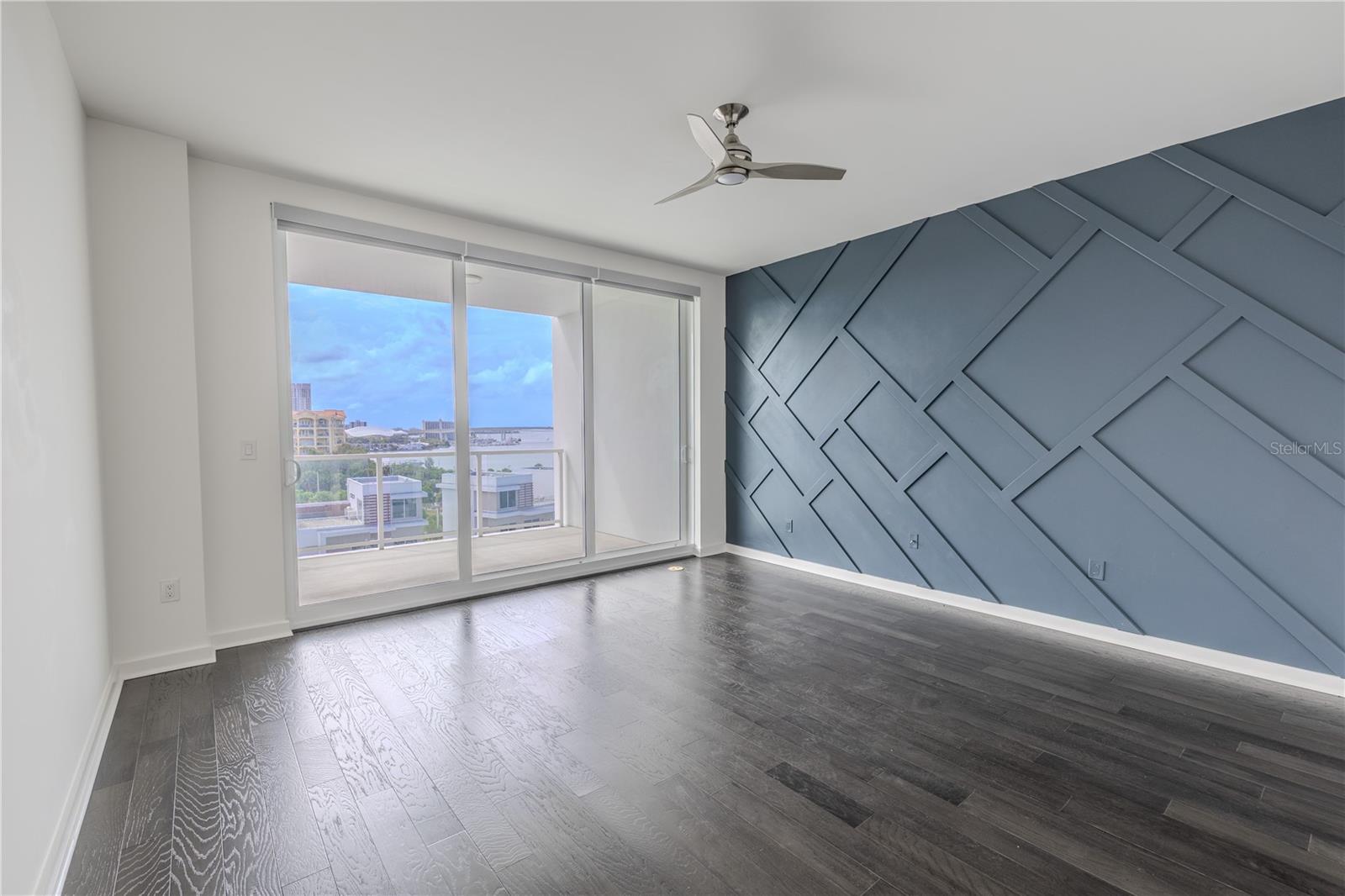 The primary bedroom features a striking custom accent wall that adds architectural depth and modern sophistication. Painted in a rich slate blue, the wall showcases a geometric grid pattern of raised molding, creating a dramatic focal point that complements the room’s sleek aesthetic. The textured design plays beautifully against the dark hardwood floors and natural light, offering the perfect balance of contemporary style and visual interest.