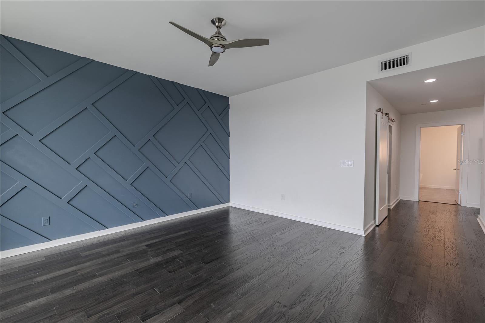The spacious primary bedroom exudes modern elegance, highlighted by a bold slate-blue geometric accent wall and rich hardwood flooring.