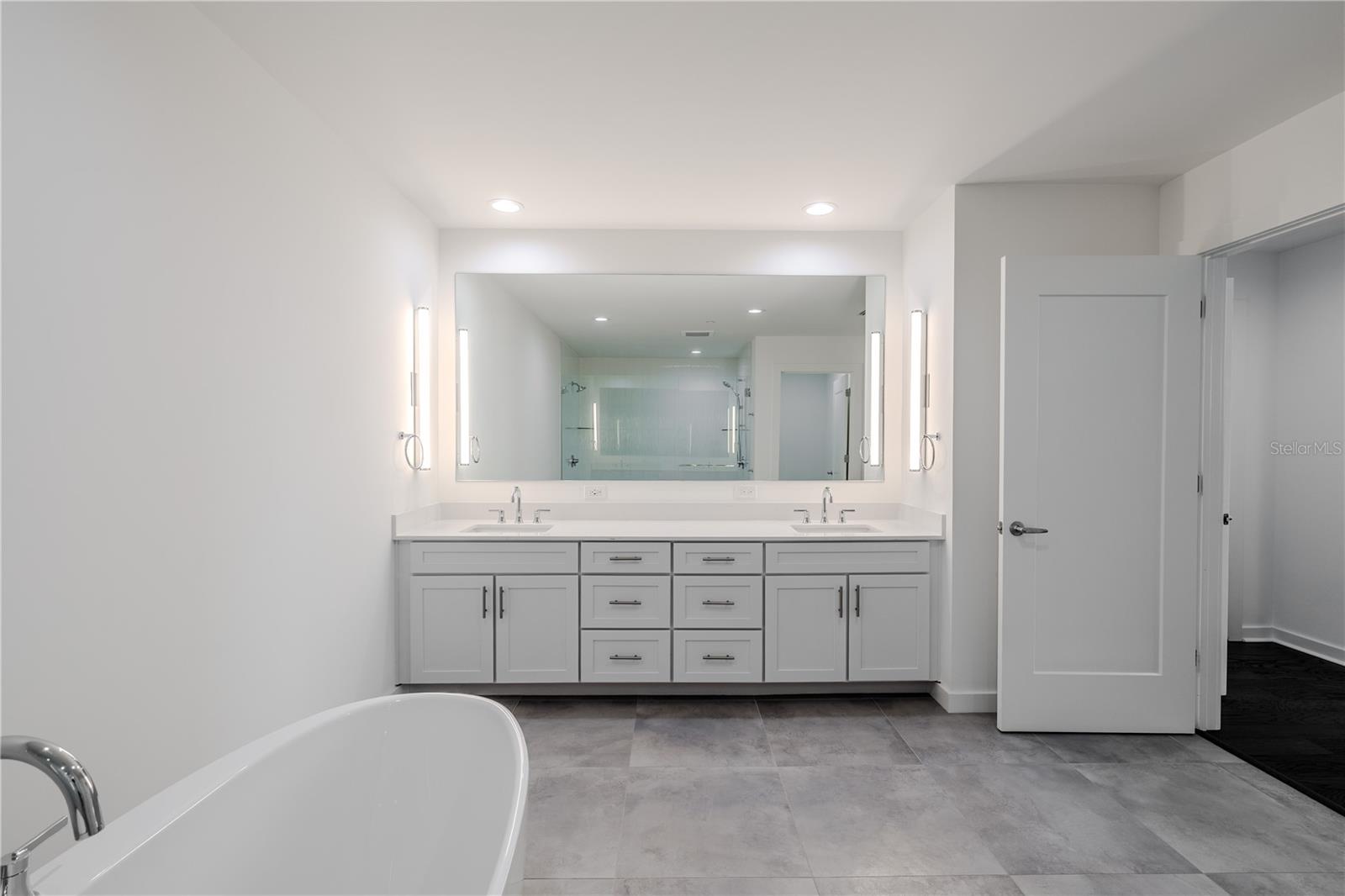 The primary bath is designed for both elegance and function, featuring a double vanity with quartz countertops, undermount sinks, and sleek chrome fixtures. Contemporary sconces flank a full-wall mirror, providing ideal lighting.