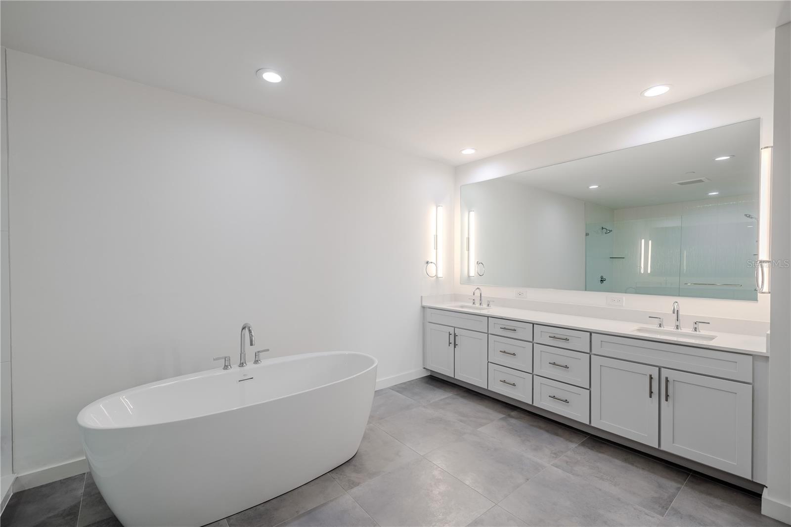 The primary bathroom features a sophisticated double vanity with an expansive quartz countertop, dual undermount sinks, and abundant drawer storage. Sleek brushed nickel hardware and soft-close cabinetry complement the clean, modern aesthetic. A full-width mirror framed by contemporary vertical sconces provides excellent lighting and adds to the spa-like ambiance of this beautifully designed space.