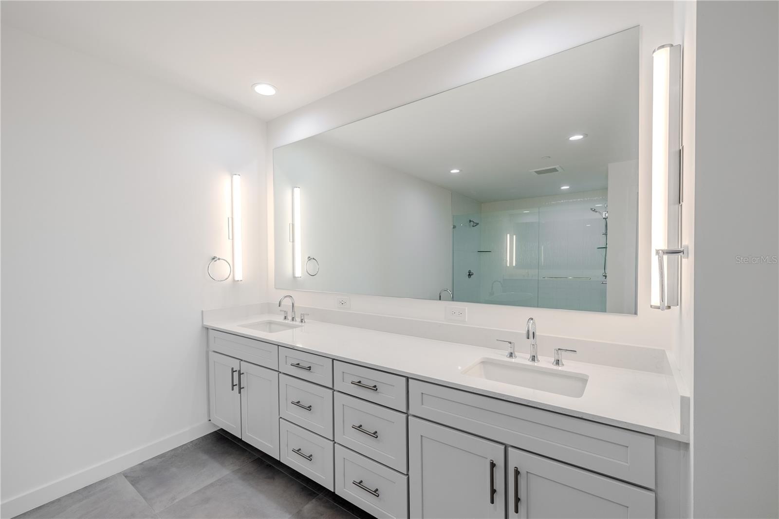 This elegant double vanity features quartz countertops, dual undermount sinks, and sleek chrome fixtures. The expansive mirror is beautifully framed by modern vertical sconces, offering perfect lighting for daily routines. Ample drawer and cabinet storage keep the space both stylish and functional, while the clean lines and neutral palette enhance the bathroom’s serene, spa-like atmosphere.