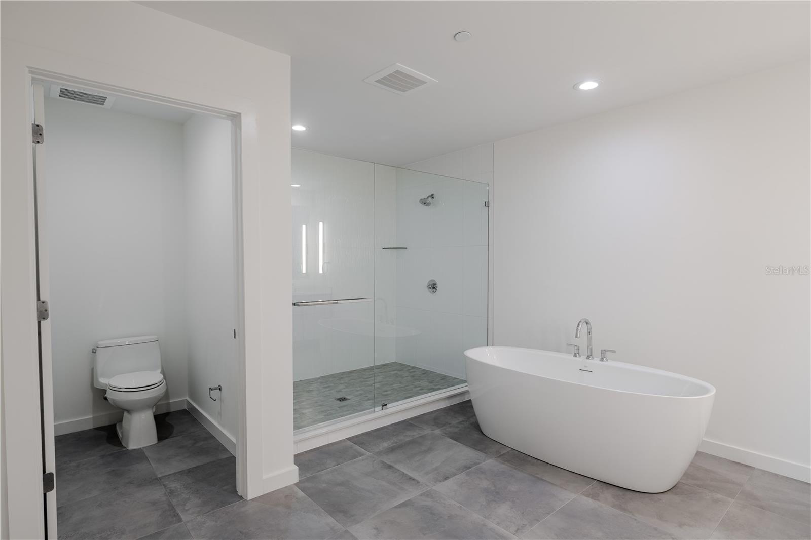 The luxurious primary bathroom exudes spa-like serenity with a sleek, modern design. A private water closet, a freestanding soaking tub serves as the elegant focal point, complemented by a frameless glass-enclosed walk-in shower