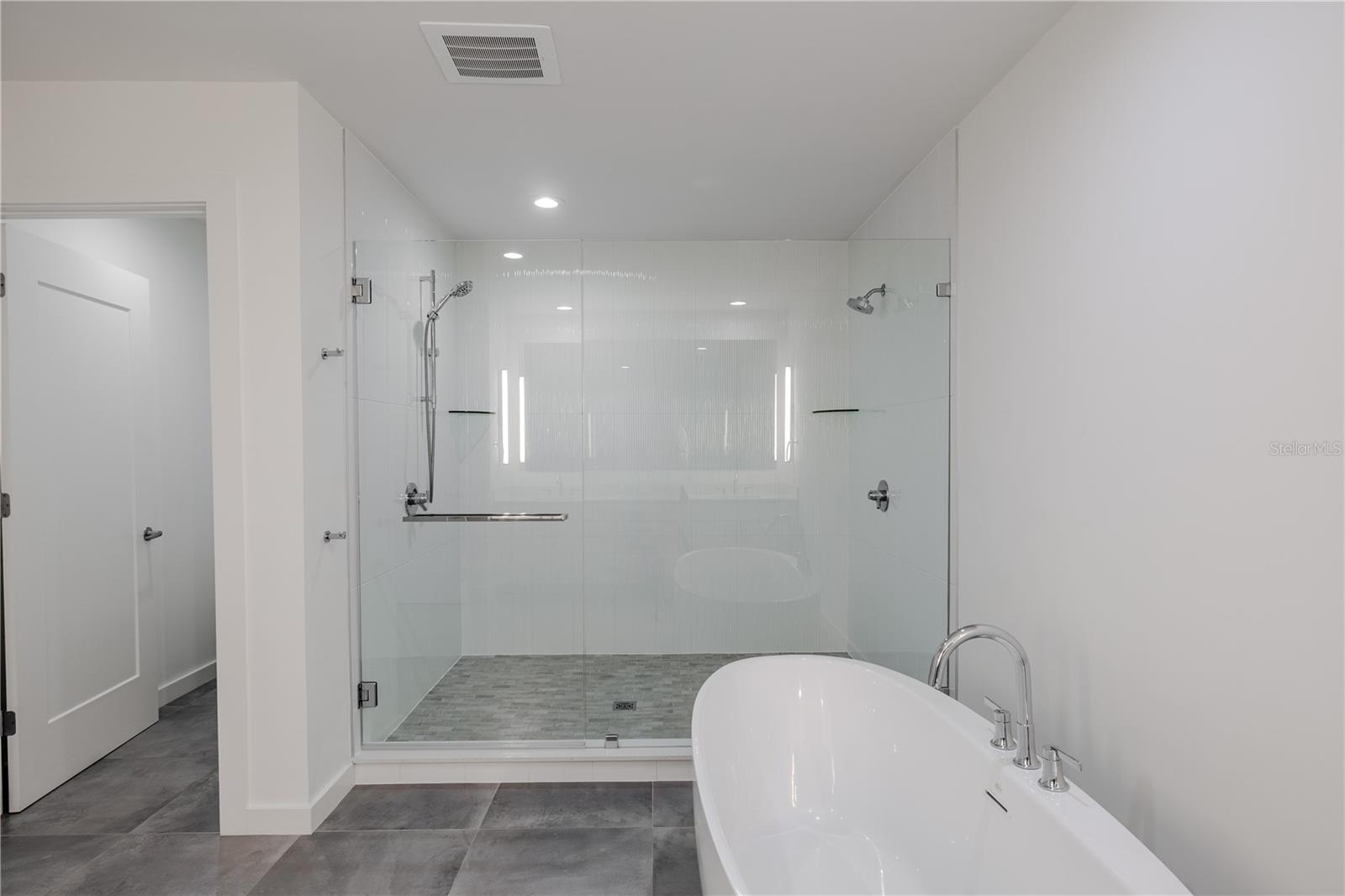 The primary bath showcases a frameless glass walk-in shower featuring dual shower heads, sleek chrome fixtures, and built-in shelving for a minimalist, modern look. The elegant freestanding soaking tub sits gracefully beside the shower, creating a luxurious retreat perfect for unwinding. Subtle textured wall tile and large floor tiles enhance the clean, contemporary aesthetic of this serene space.