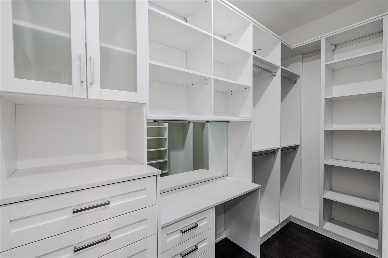 The secondary primary closet offers exceptional organization and functionality, featuring custom built-ins with open shelving, drawers, and hanging space. Frosted glass cabinet doors add a touch of elegance, while a built-in vanity with mirror and ample countertop space provides a perfect spot for getting ready. Thoughtfully designed to maximize storage and style