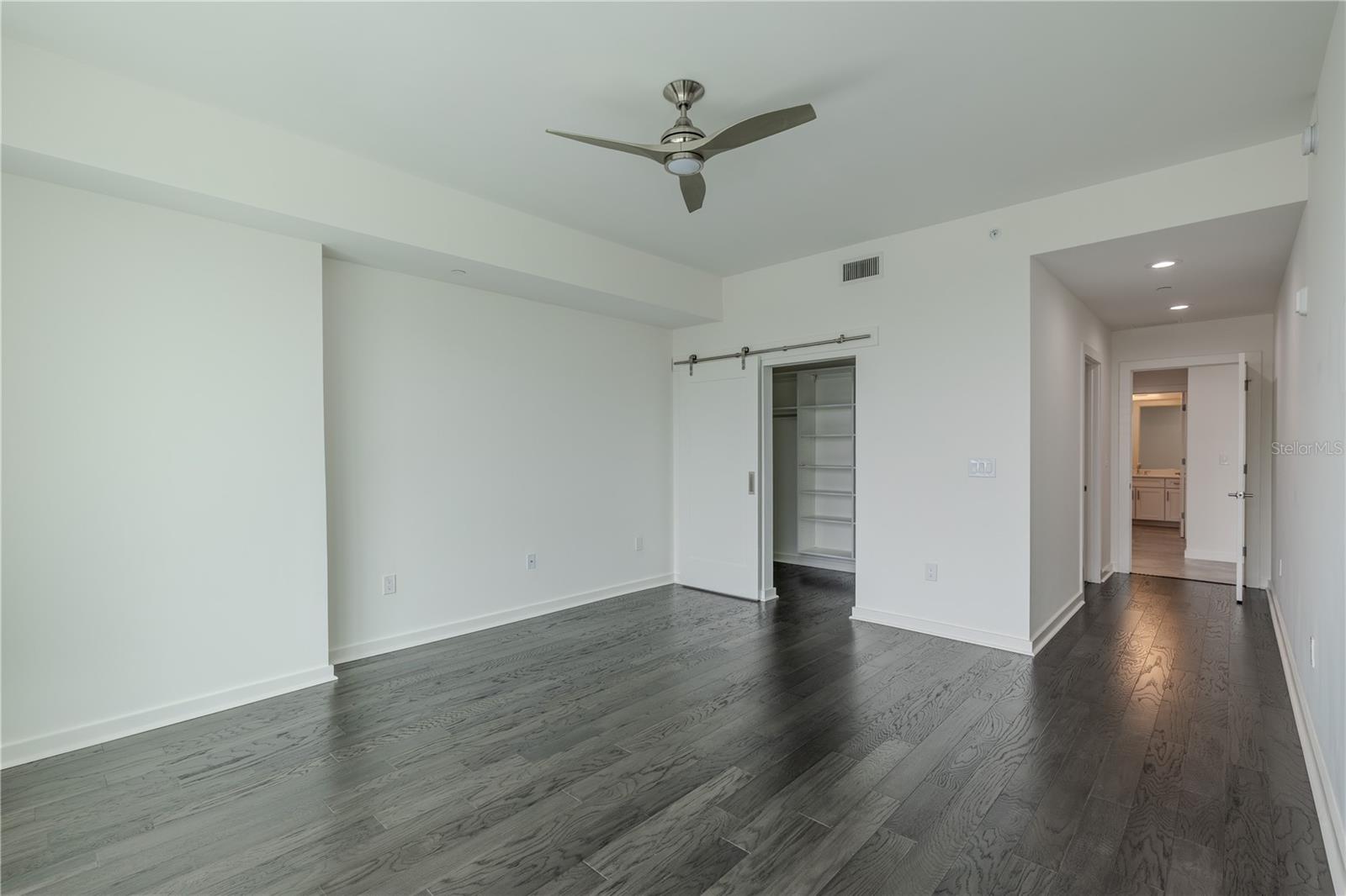 This inviting second bedroom features rich hardwood floors, recessed lighting, and a sleek ceiling fan that adds a touch of modern style. A sliding barn door opens to a custom closet with built-in shelving for organized storage. The room connects conveniently to an en-suite bath
