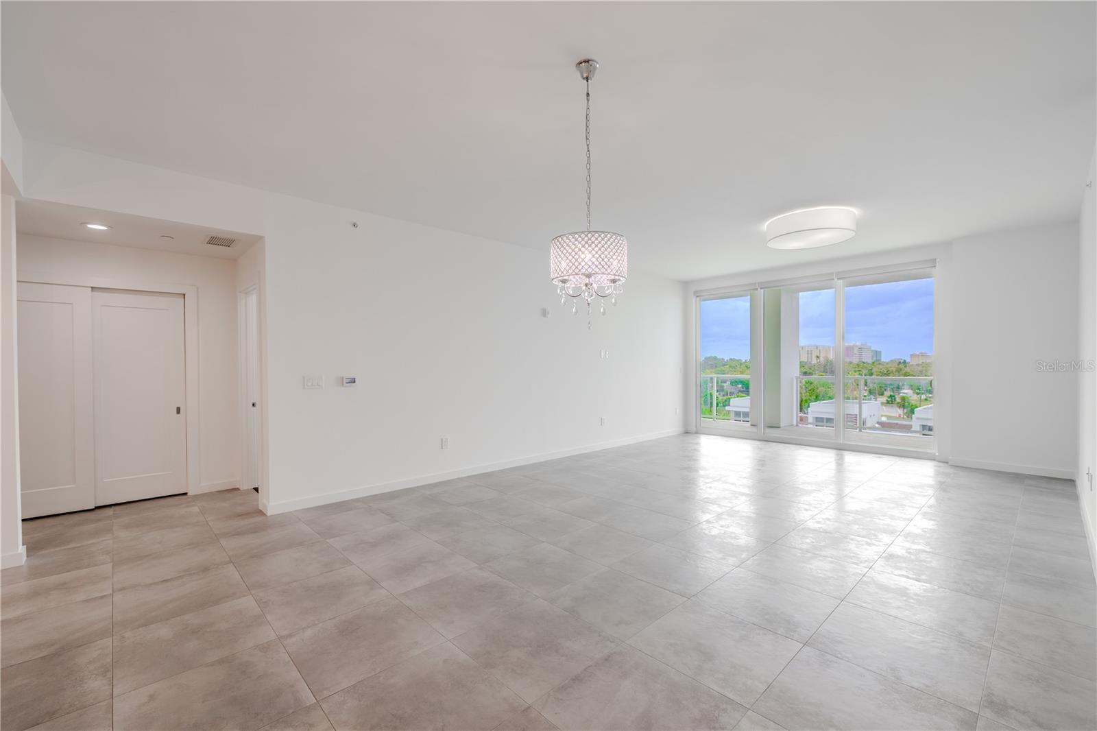 This spacious open-concept dining and living area is bright and contemporary, featuring large-format porcelain tile floors and floor-to-ceiling sliding glass doors that fill the space with natural light. A sparkling crystal chandelier defines the dining area, while the adjoining living space offers seamless access to the balcony and stunning views of the Intracoastal and city skyline. The clean, neutral palette provides the perfect backdrop for any design style.
