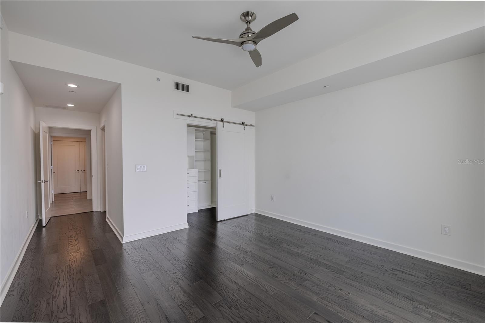 Bedroom 3 offers a sleek and versatile layout enhanced by dark hardwood floors, crisp white walls, and clean architectural lines. A modern ceiling fan adds comfort and style, while the hallway leads seamlessly to the en-suite bath and custom closet.