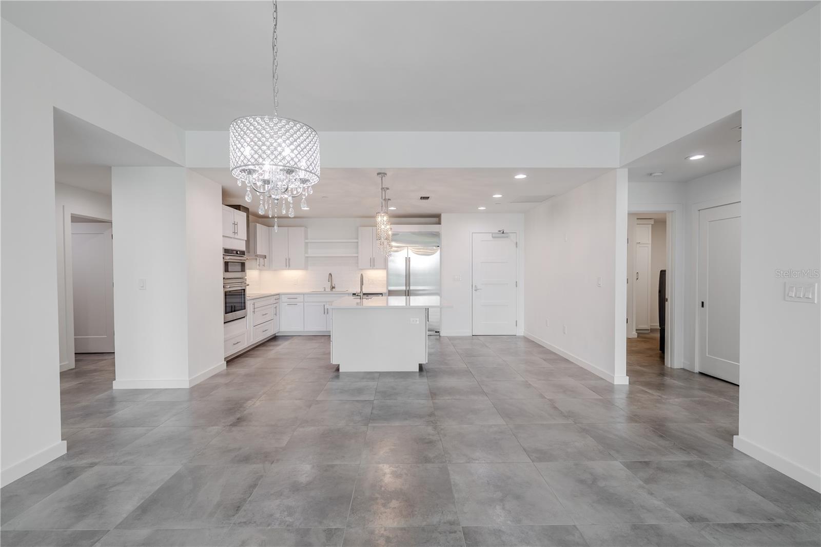 Open, split floor plan with a clear view into the modern kitchen featuring a large center island, stainless steel appliances, and white shaker cabinetry. Contemporary lighting and sleek gray tile flooring create a bright, cohesive flow throughout the main living area.