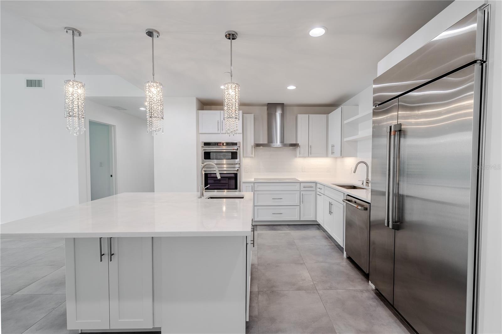 This stunning modern kitchen combines sleek design with high-end functionality. A spacious quartz-topped island anchors the open layout, offering bar seating and a built-in sink beneath a trio of sparkling crystal pendant lights.
