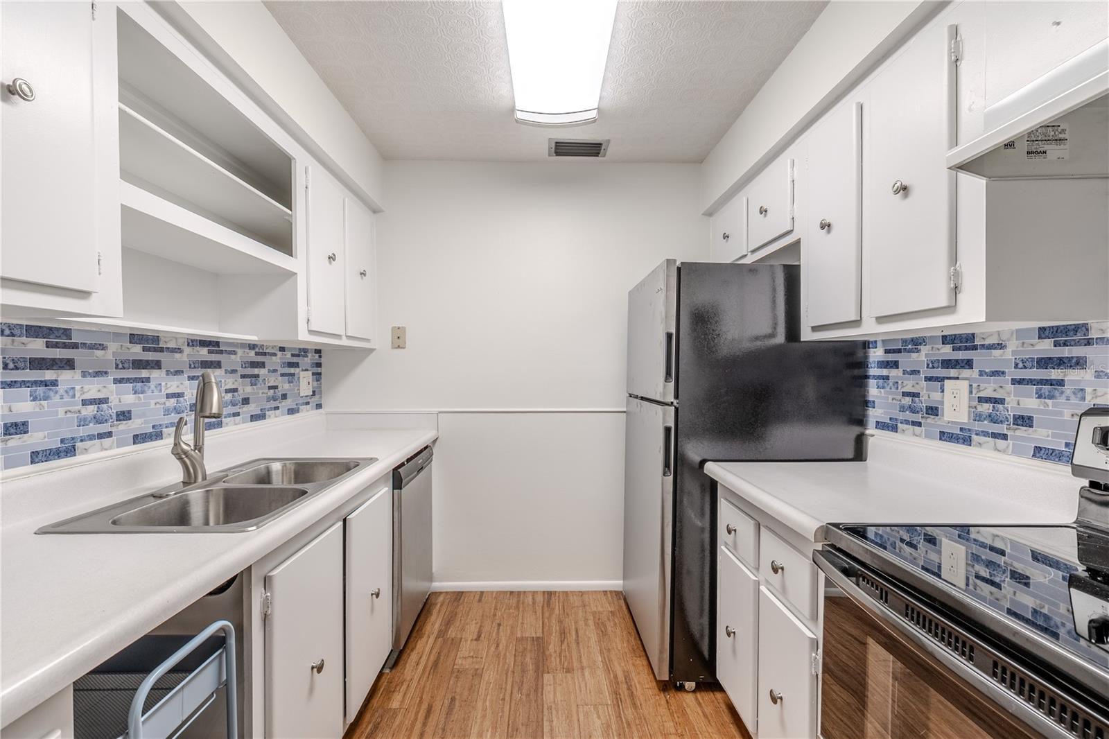 The kitchen features a clean, functional layout with crisp white cabinetry, stainless steel appliances, and a double sink for added convenience. The blue mosaic-style backsplash adds a fresh coastal touch and beautifully complements the warm bamboo flooring. Ample counter space and open shelving provide both practicality and charm, making meal prep and storage easy.