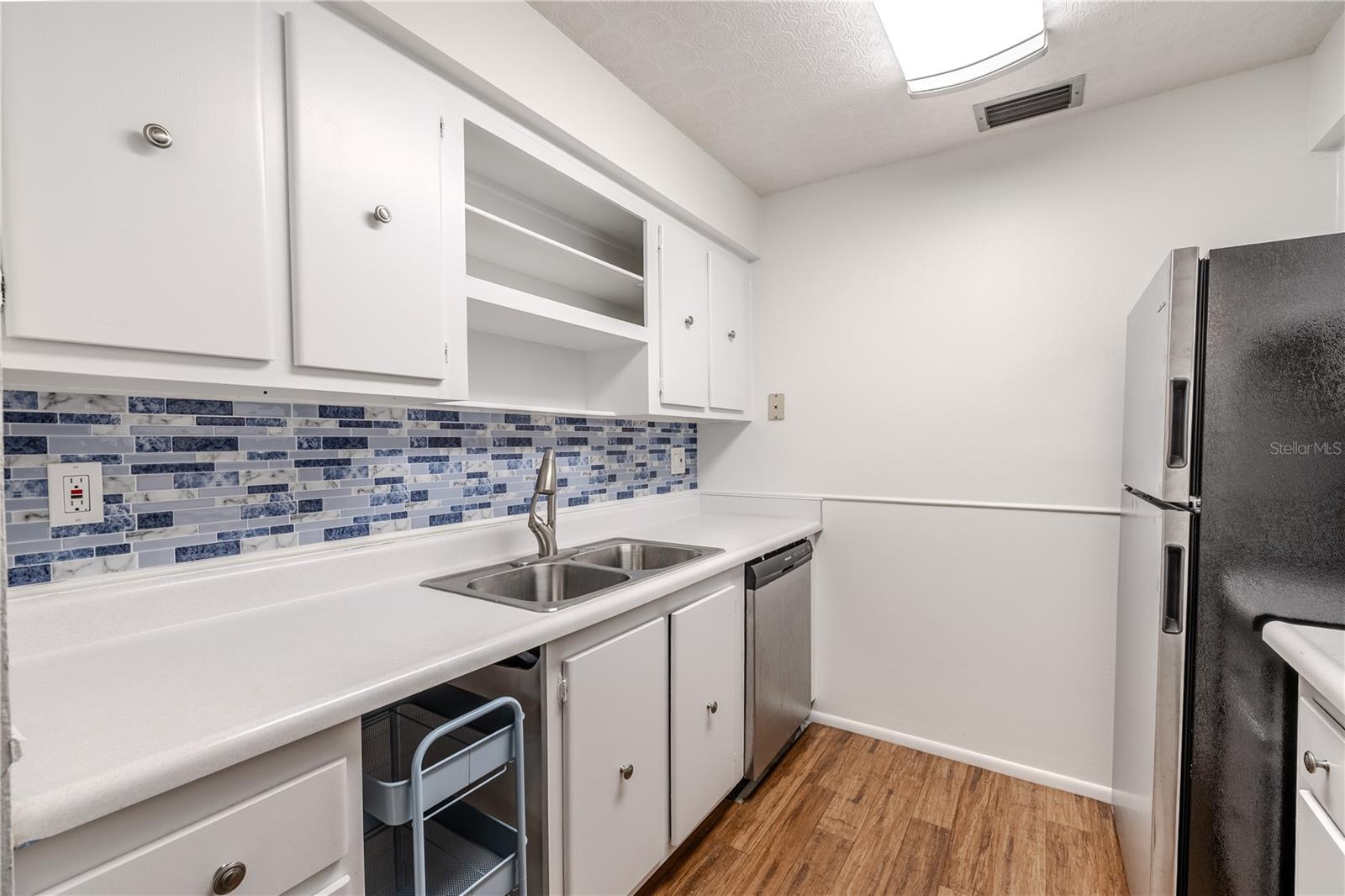 This bright and efficient kitchen features classic white cabinetry with ample storage and a clean, streamlined design. The double stainless steel sink and brushed nickel faucet add modern functionality. Open shelving offers easy access to everyday essentials or decorative display, and the stainless steel dishwasher provides added convenience.