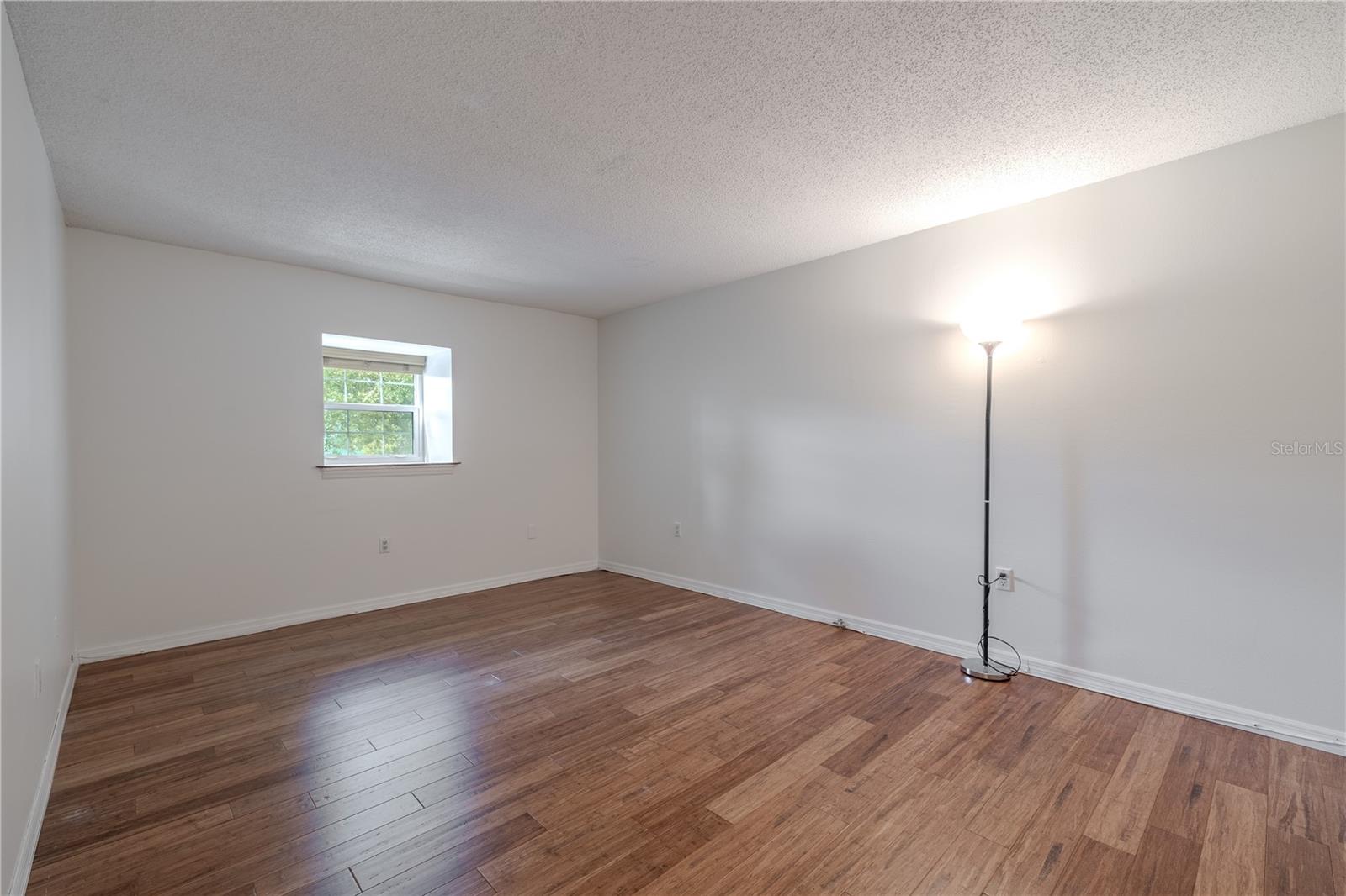 The primary bedroom is spacious and serene, with warm bamboo flooring and soft neutral walls that create a calming atmosphere. A large double-pane window fills the room with natural light and provides a pleasant view of the surrounding greenery