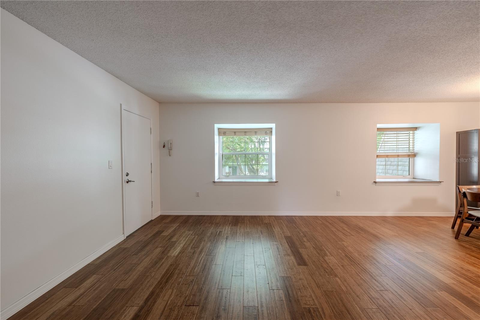 The living room features sleek bamboo flooring that adds warmth and natural elegance to the space. Neutral walls and a textured ceiling create a clean, airy atmosphere, while two large windows fill the room with natural light.