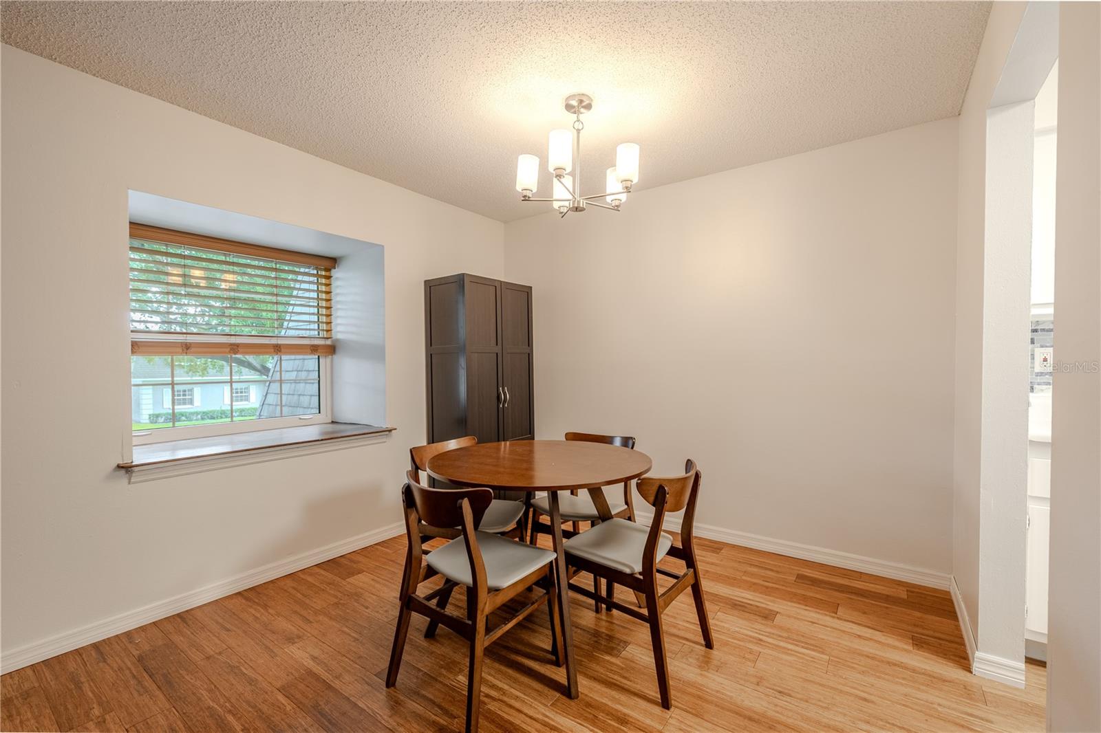 The dining room offers a cozy and inviting space with warm bamboo flooring and soft neutral walls that create a bright, modern feel. A large double pane window with wood blinds fills the area with natural light, providing a pleasant view of the tree-lined community. The contemporary light fixture adds a touch of elegance, while the open layout allows easy access to the adjoining kitchen—perfect for entertaining or casual meals. This space comfortably accommodates a dining set