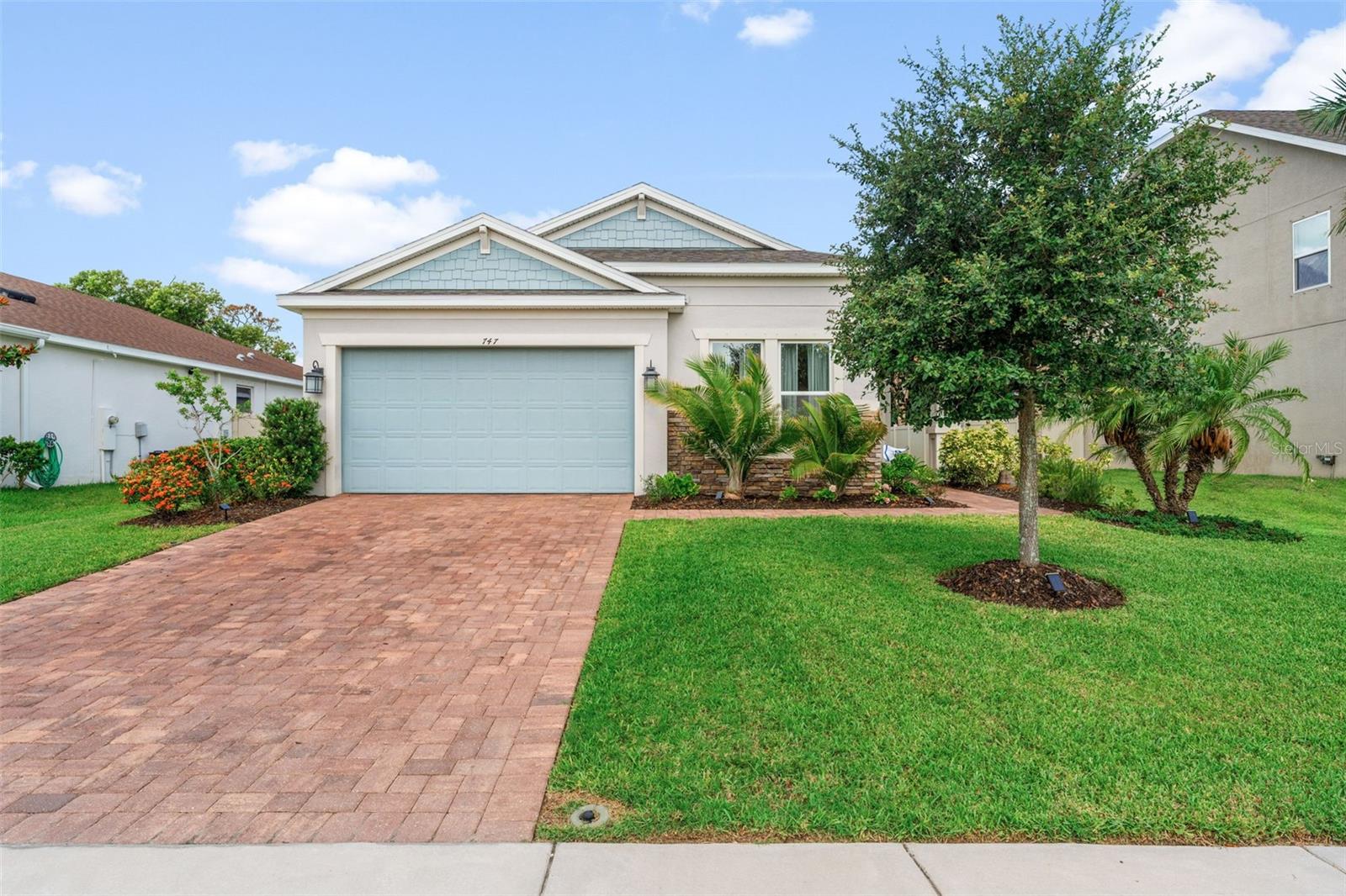 Beautifully maintained 2018-built home with coastal-inspired architecture, stone accents, and a brick paver driveway leading to a two-car garage. Lush tropical landscaping adds curb appeal and warmth.