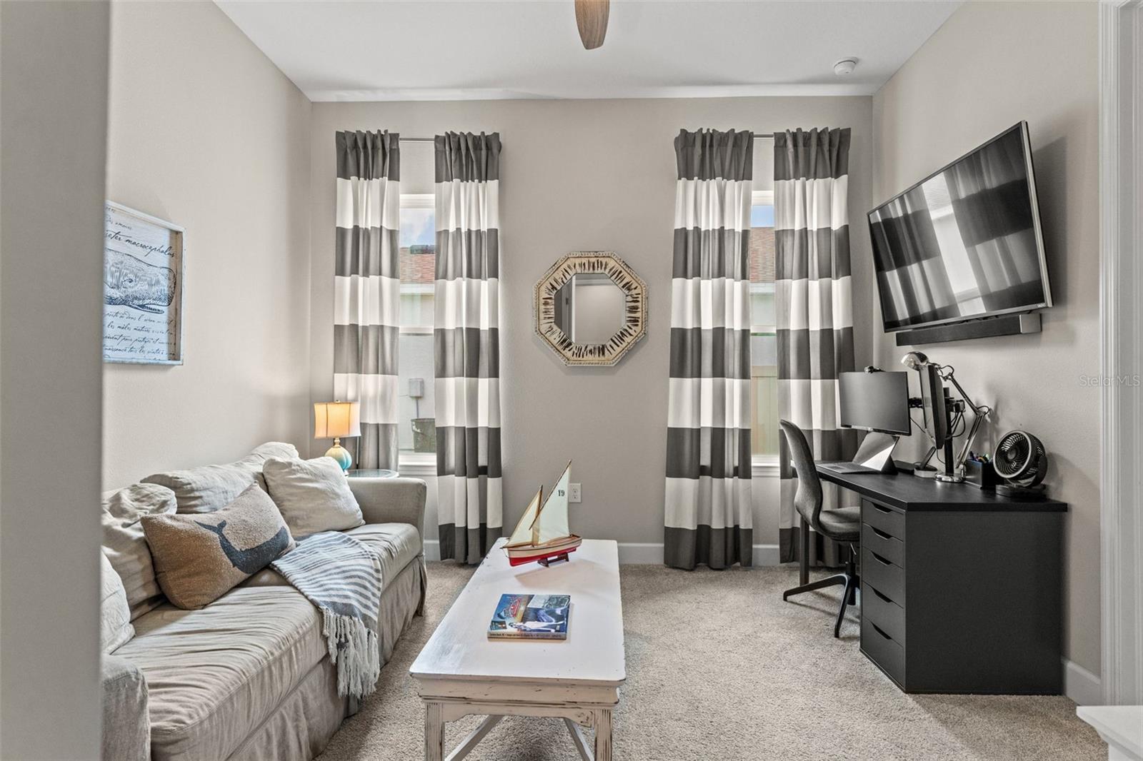 Flexible guest suite currently used as a home office and lounge, featuring large windows, soft natural light, and custom drapery.