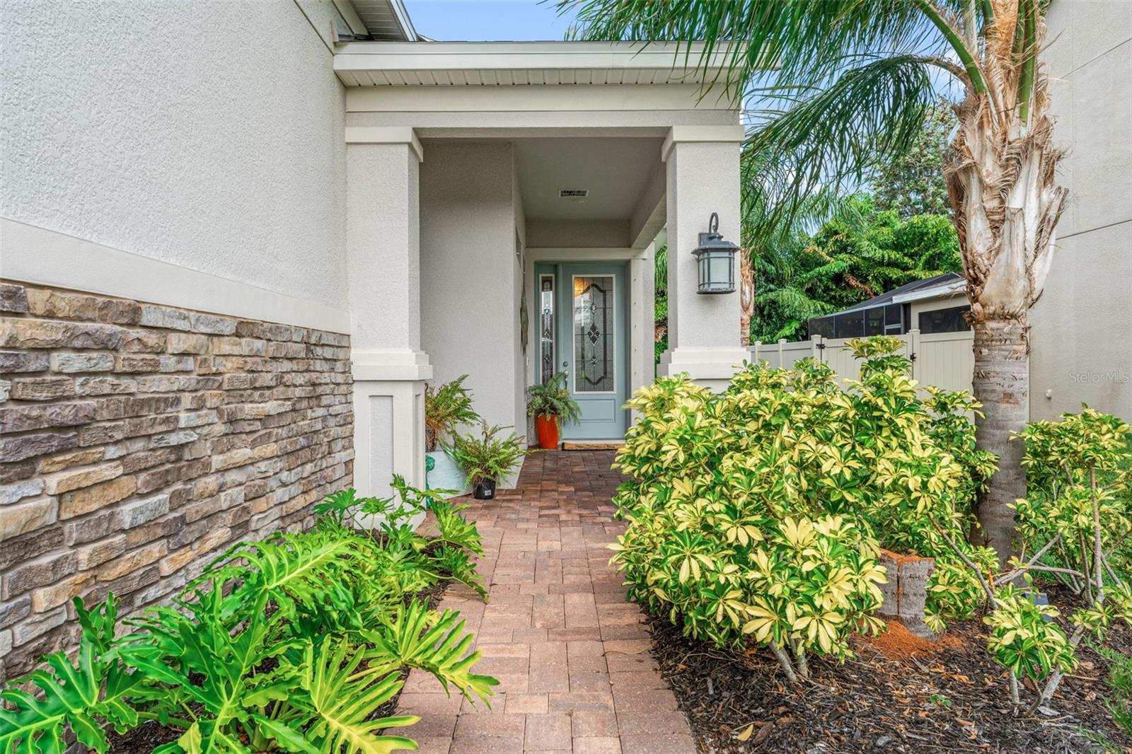 Brick-paver walkway framed by vibrant landscaping leads to a covered entry with stone detailing and an upgraded glass door for a welcoming first impression.