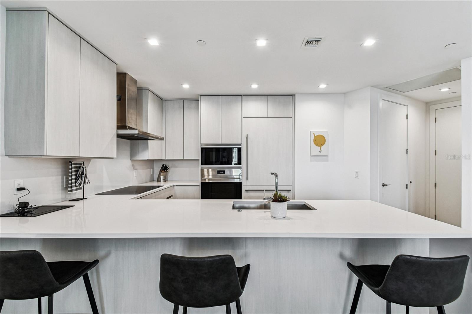 Sleek, contemporary kitchen featuring a large quartz peninsula with seating, paneled refrigerator, wall oven/microwave, induction cooktop, and streamlined cabinetry with recessed lighting.