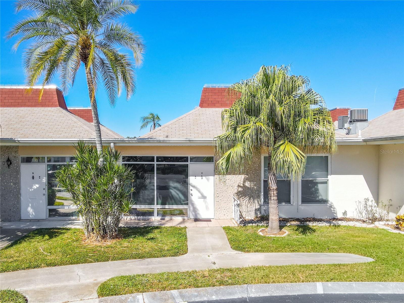 A welcoming condo entrance features a neatly paved sidewalk lined with tall, graceful palm trees on either side, creating a tropical and inviting approach. The pathway leads up to a sleek storm door that offers both style and protection, accented by a transom window above that allows natural light to filter into the entryway. The combination of greenery and clean architectural details gives the entrance a bright, resort-like charm while maintaining a sense of island elegance.