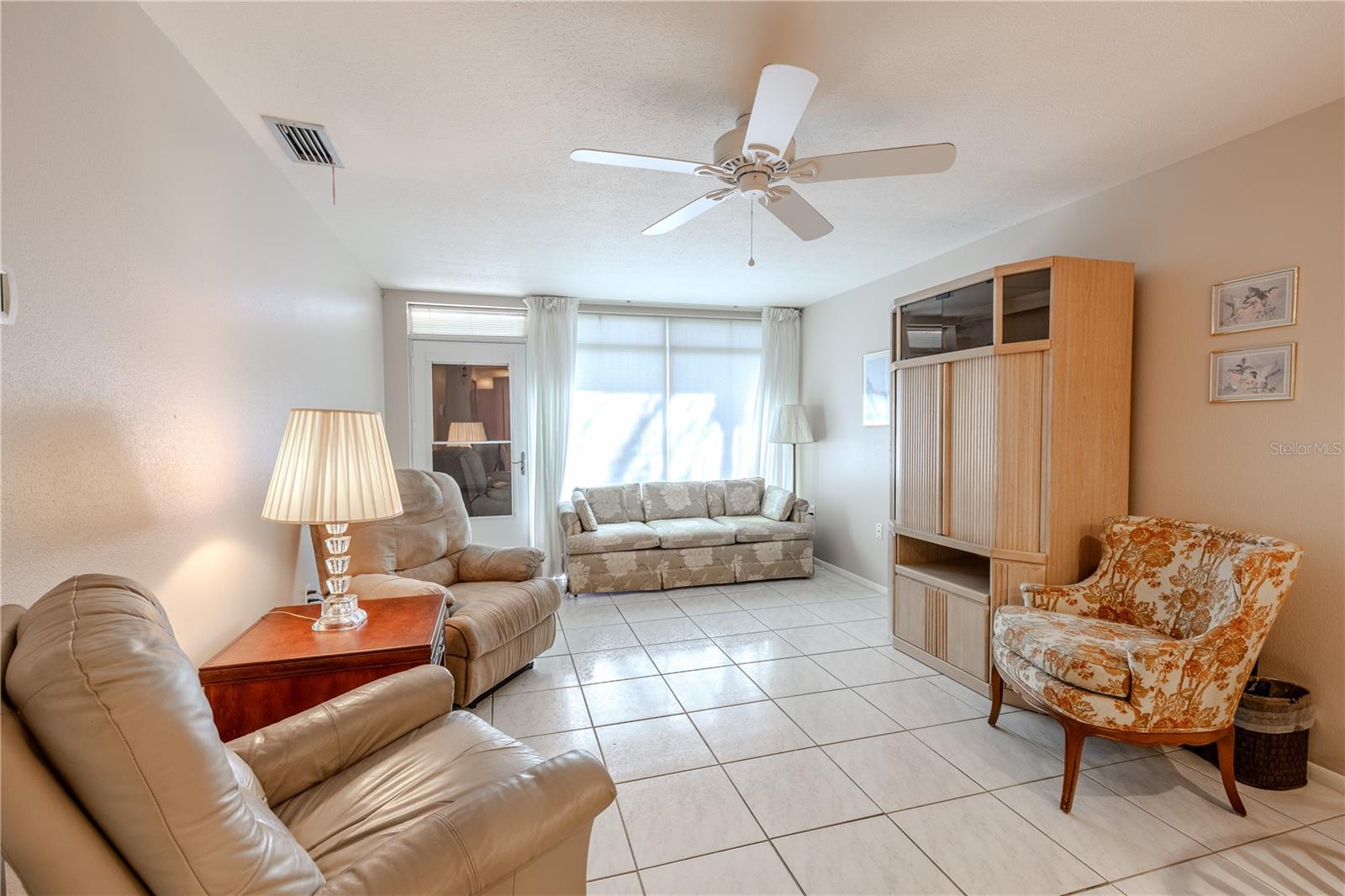 View of the living room from the opposite side, showcasing the airy, open layout that connects seamlessly to the kitchen. Large front windows fill the space with natural light, while the ceiling fan ensures comfort. The main entrance and interior two-paneled glass door, visible when the storm door is open, add charm and functionality, highlighting the bright, welcoming atmosphere and smooth flow throughout the home.