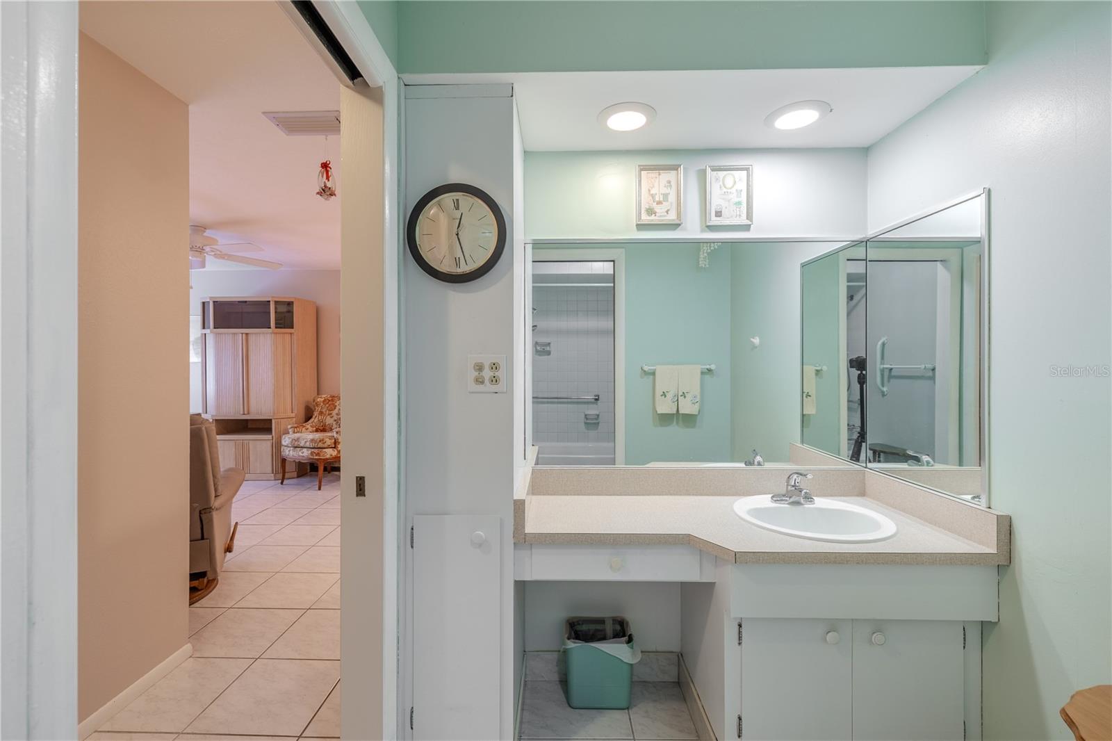 Everyday ease shines in this bathroom set just off the main living area. A sit-down vanity with extended counter space and a wide wrap-around mirror under recessed lighting makes getting ready comfortable and unhurried. The step-in shower with a grab bar adds peace of mind, while towels are within easy reach. Tile floors keep cleanup simple and connect smoothly to the adjoining room, so your morning routine flows naturally from coffee to “out the door.”