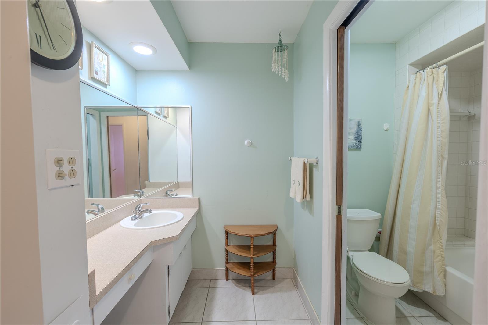Bright and practical, this bathroom is organized for smooth daily routines. An angled single-sink vanity offers generous counter space, a wide wrap-around mirror, and recessed lighting—great for getting ready without feeling cramped. Tile floors and soft sea-glass walls keep the space fresh and easy to maintain. To the right, a separate alcove with the tub/shower and toilet adds privacy, so one person can shower while another uses the vanity. Thoughtful touches—a towel bar within reach and a small corner shelf for essentials—make this a calm, efficient spot from morning to bedtime.