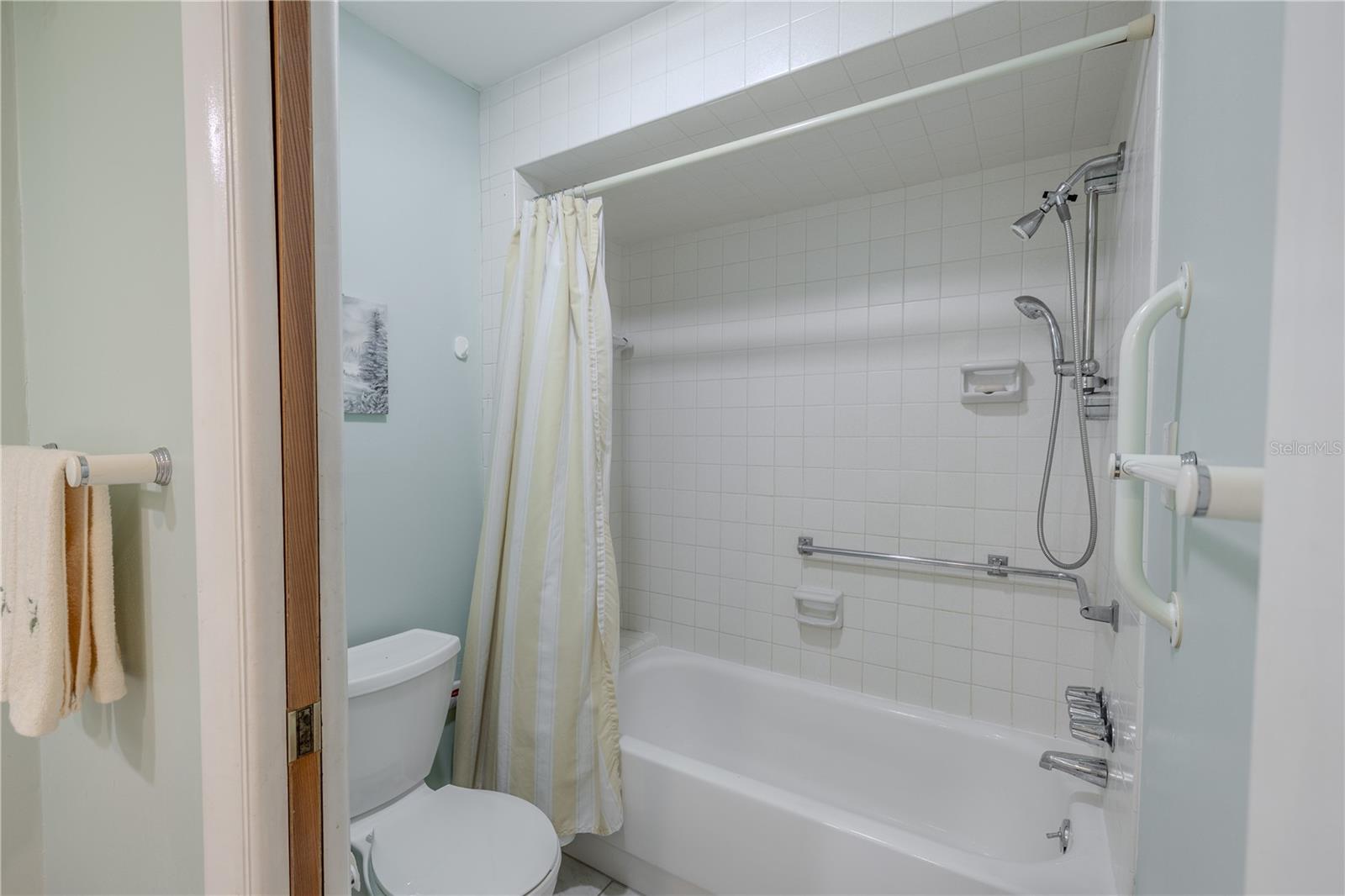 Bright and simple tub/shower combo with a clean white tile surround, overhead shower head plus a hand held shower, and multiple grab bars for added confidence when bathing. A neutral shower curtain, soft sea-glass wall color, and nearby towel bar keep the space calm and practical. With the toilet tucked beside the tub and everything within easy reach, this bath is set up for quick morning showers or an unhurried soak at day’s end—low-maintenance and comfortable.