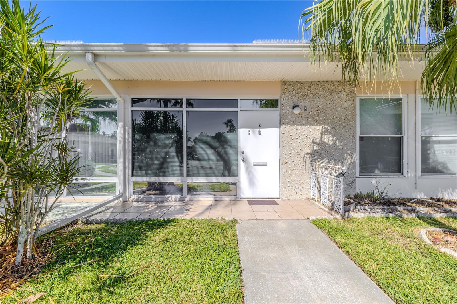A welcoming condo entrance features a paved sidewalk revealing the wide pathway leading up to the entrance accented by expansive windows and a protective storm door, with a transom window above that allows natural light to filter into the entryway.