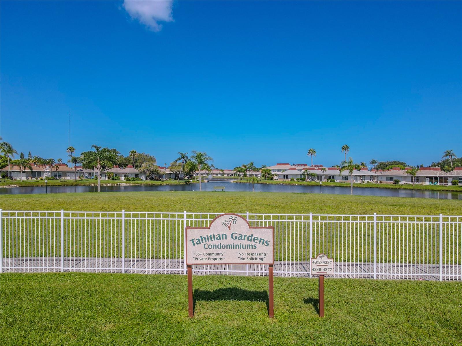 Charming ground-level view of the Tahitian Gardens Condominiums entrance sign, gracefully framed by a white wrought-iron fence. Behind it, a tranquil pond and expansive green lawns create a serene, park-like setting, welcoming residents and visitors into a peaceful, beautifully landscaped community. The scene captures the inviting curb appeal and relaxing ambiance that define life at Tahitian Gardens.