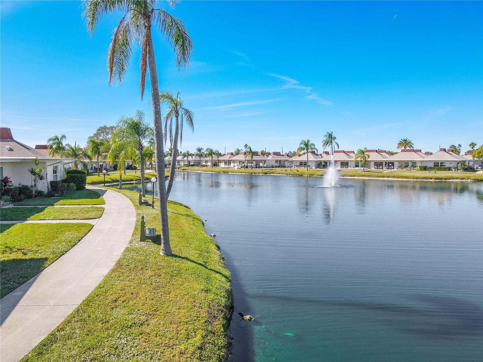 Scenic walking path winding around a serene, palm-lined pond with a beautiful fountain as the focal point in the background. The pathway offers a peaceful setting for leisurely strolls, surrounded by lush landscaping and tranquil water views.