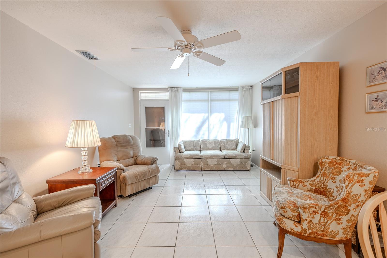 View of the full living room from the kitchen, where sunlight streams through large front windows, creating a bright and welcoming atmosphere. A ceiling fan adds comfort, while the main entrance and interior two-paneled glass door, visible when the storm door is open, enhance the airy, open feel of the space. This layout offers a seamless connection between rooms, perfect for relaxing, entertaining, or enjoying daily life in a light-filled, inviting home.