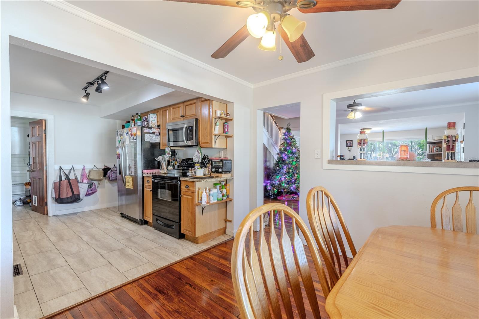 This view from the far side of the dining room beautifully showcases the open and connected layout of the main living areas. The dining room seamlessly transitions into the kitchen, complete with tile flooring, wood cabinetry, and modern appliances, while the wide passthrough and adjoining doorway offer clear sightlines into the spacious living room. Original wood flooring continues throughout the dining and living spaces, creating warmth and cohesion. This open-concept flow enhances both everyday convenience and the home’s welcoming atmosphere, making it ideal for gatherings and family living.
