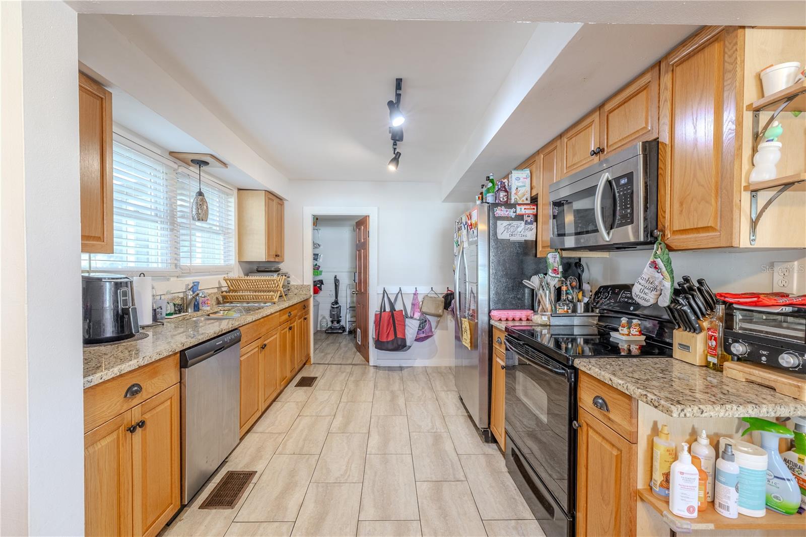 This bright and functional galley-style kitchen features warm wood cabinetry, granite countertops, and modern stainless-steel appliances. Large windows along the left wall fill the space with natural light, creating an inviting environment for cooking and gathering. The tile flooring adds durability and easy maintenance, while overhead track lighting ensures excellent visibility. At the far end of the kitchen, a walk-in pantry offers exceptional storage space and convenience—perfect for keeping everything organized and within reach. This well-designed kitchen combines practicality with comfort, making it a standout feature of the home.