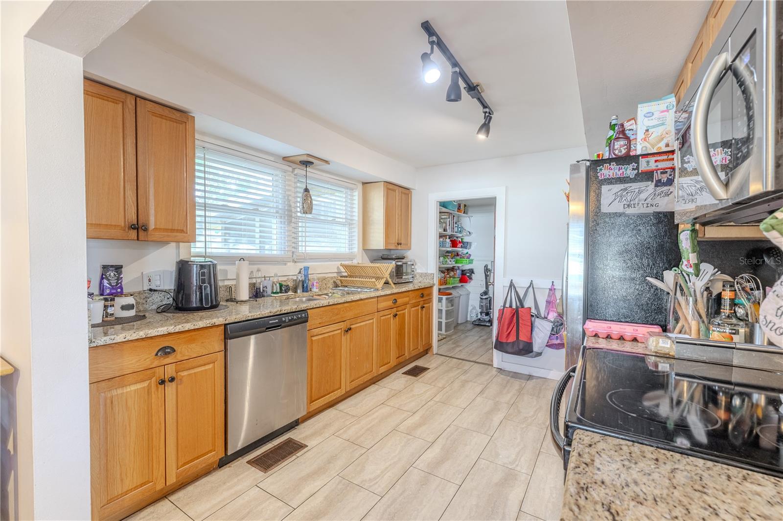 This opposite angle of the kitchen highlights the long stretch of granite countertops, abundant cabinetry, and stainless-steel dishwasher beneath a bank of bright windows. The natural light pours in from the large window overlooking the expansive elevated wooden deck, offering a pleasant view while cooking or entertaining. Tile flooring adds durability, and track lighting provides excellent illumination throughout the space. At the far end, the doorway leads to the spacious walk-in pantry, giving the kitchen both convenience and functionality.