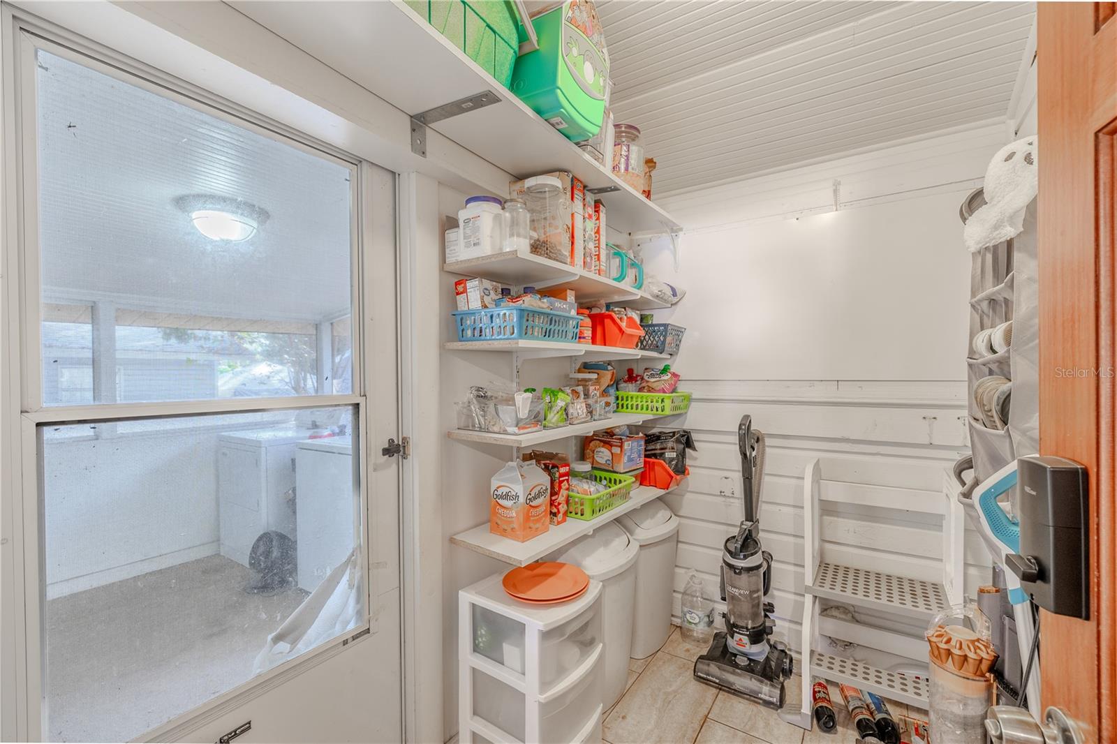 The spacious walk-in pantry offers excellent storage with multiple built-in shelves and room for bulk items and household essentials. To the left, a door opens directly to the screened-in porch, where the washer and dryer are conveniently located—making laundry easily accessible while keeping it separate from the main living areas. This combined pantry and utility setup adds valuable functionality and efficiency to the home’s kitchen layout.