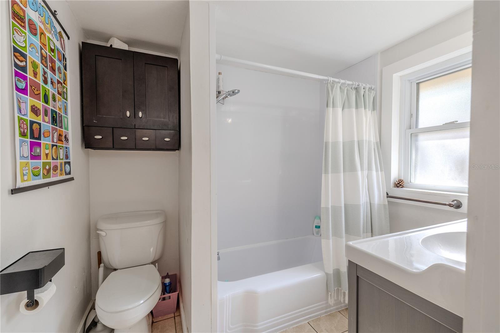 This first-floor en- suite full bathroom offers convenience and functionality, featuring a combination shower and tub, a modern vanity, and a bright window that brings in natural light. A wall-mounted cabinet above the toilet provides additional storage. This bathroom is accessible directly from the first-floor bedroom as well as from the kitchen, making it ideal for guests, everyday use, or multigenerational living.