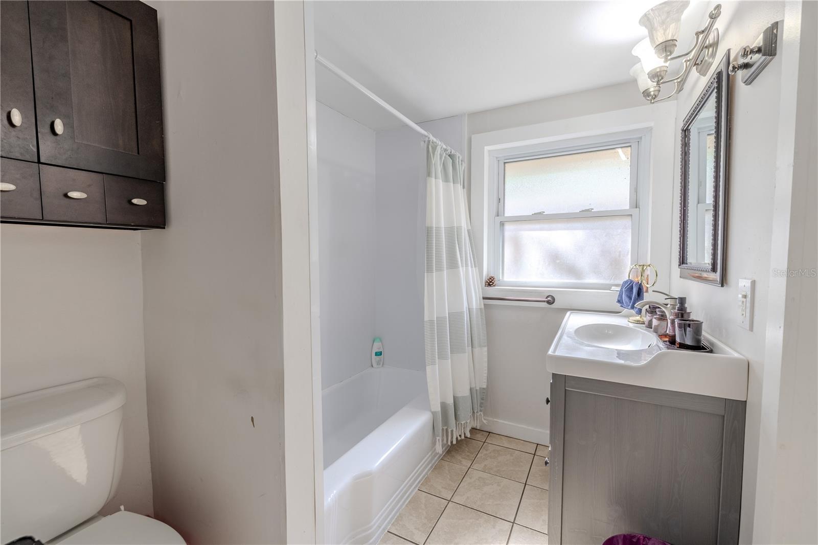 This additional view of the first-floor en-suite bathroom highlights the large frosted window that fills the space with soft natural light while maintaining privacy. The room features a combination shower and tub, a modern vanity, and neutral tile flooring. With access from both the first-floor bedroom and the kitchen, this bright and functional bathroom adds convenience for guests and everyday living.