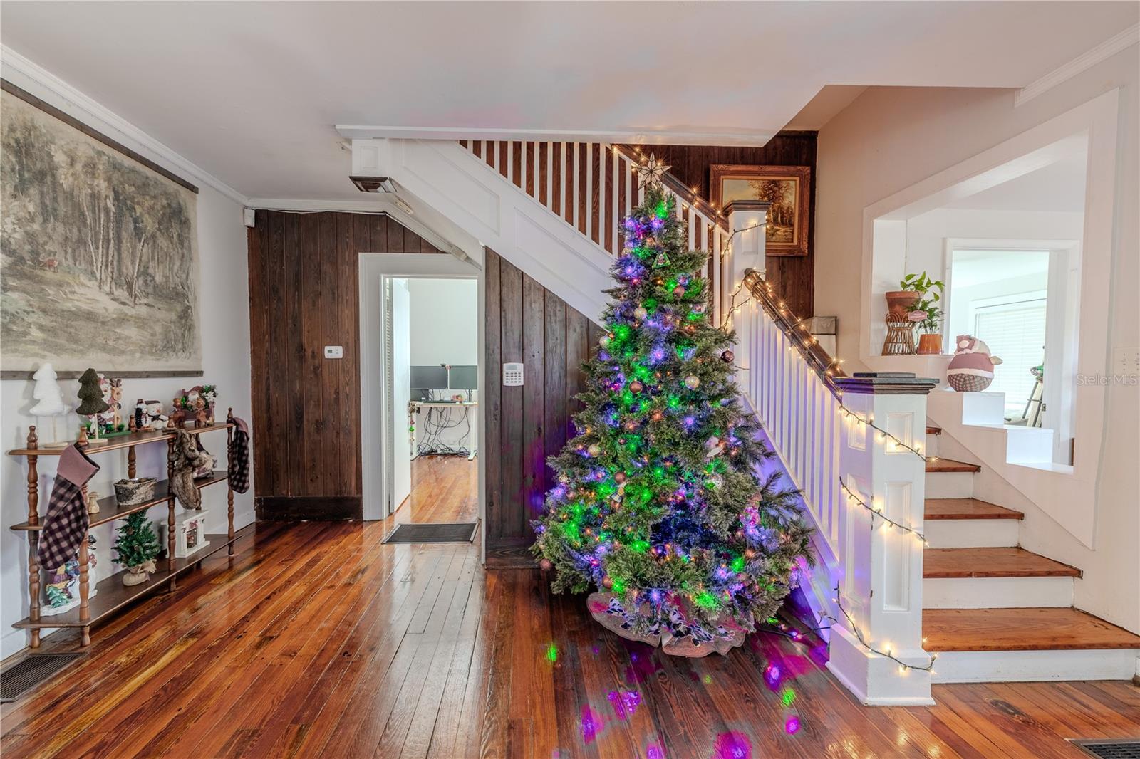 This view from the living room highlights the home’s charming staircase with its classic wood detailing and crisp white railing, adding character to the main level. Just beyond the stairs is the doorway to the first-floor bedroom, offering convenient access while maintaining privacy. The rich original wood floors and warm wood accent wall enhance the inviting feel of the space, showcasing the historic charm and functional layout of the home’s main living area.