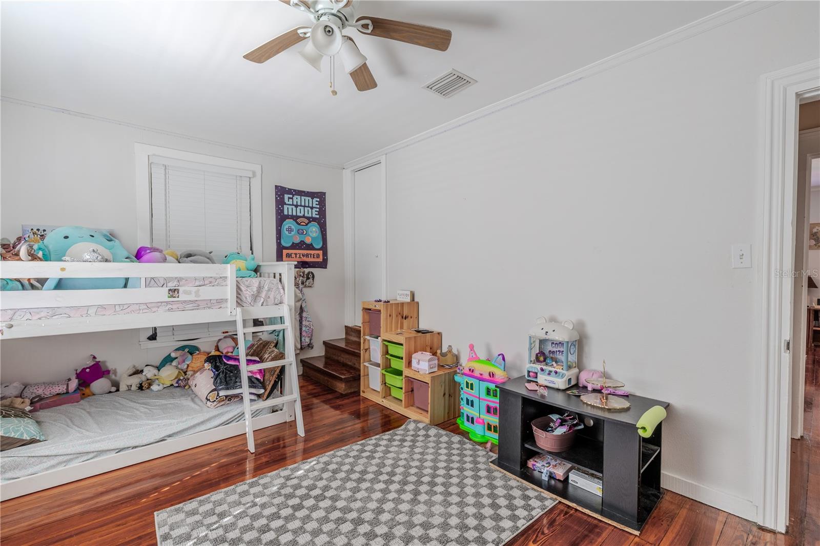 This angle of the second-floor bedroom highlights the convenient built-in corner closet, providing practical storage without taking up additional floor space. The room features gleaming wood floors, neutral walls, and a ceiling fan for added comfort. With plenty of room for furniture and play, this versatile space is ideal for a child’s bedroom, guest room, or creative area.