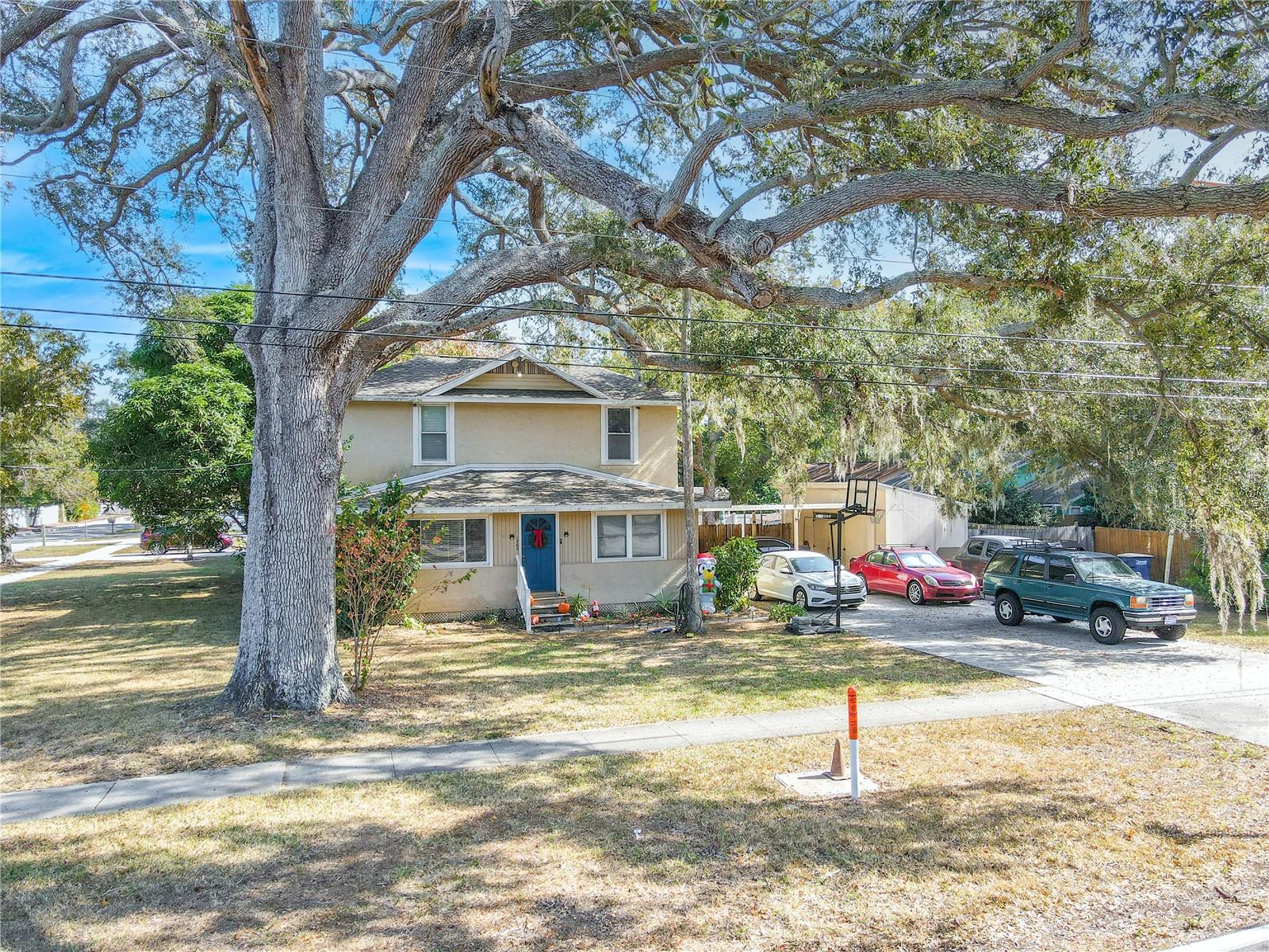 This welcoming front view showcases the home’s charming 1918 character, set beneath the sweeping branches of a magnificent mature oak. The property features a spacious front yard on a desirable corner lot, bordered by sidewalks for easy neighborhood strolls. A bold blue front door adds a touch of personality, while the extended driveway to the right provides abundant parking and leads to the carport and workshop area. This inviting perspective highlights the home’s curb appeal, shade-filled setting, and generous outdoor space.