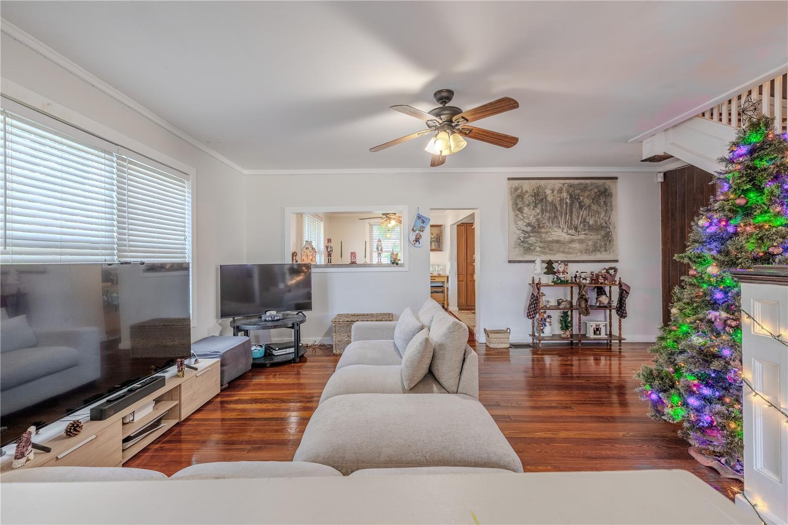 From the entry half wall, the spacious living room opens up with warm original wood floors and abundant natural light. A central ceiling fan adds comfort and airflow, complementing the room’s inviting atmosphere. This generous living area flows seamlessly toward the dining room beyond, visible through the open passthrough and doorway—creating a connected, easy-living layout perfect for gatherings and everyday living. The space offers plenty of room for seating and entertainment, all while showcasing the charm and openness of this historic home.