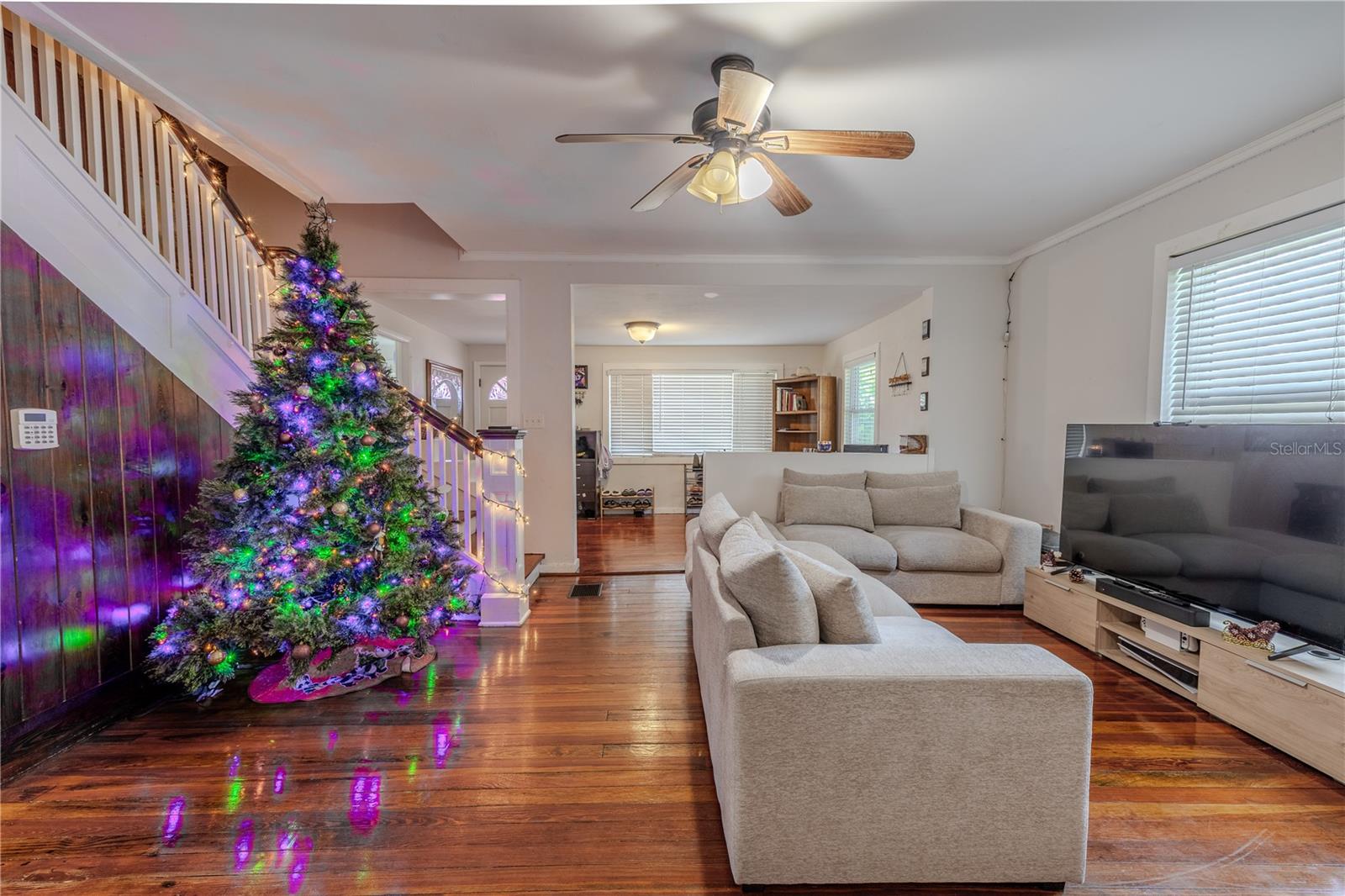 From the dining area, this view showcases the home’s expansive and connected living spaces, highlighted by beautifully preserved original wood flooring that extends throughout. The warm wood accent wall adds character and a touch of historic charm, while the open layout creates an easy flow between rooms. The staircase becomes a focal point with its classic design, and the large living room offers plenty of room for comfortable seating and everyday living. Abundant natural light and a ceiling fan enhance the inviting feel of this spacious main level.