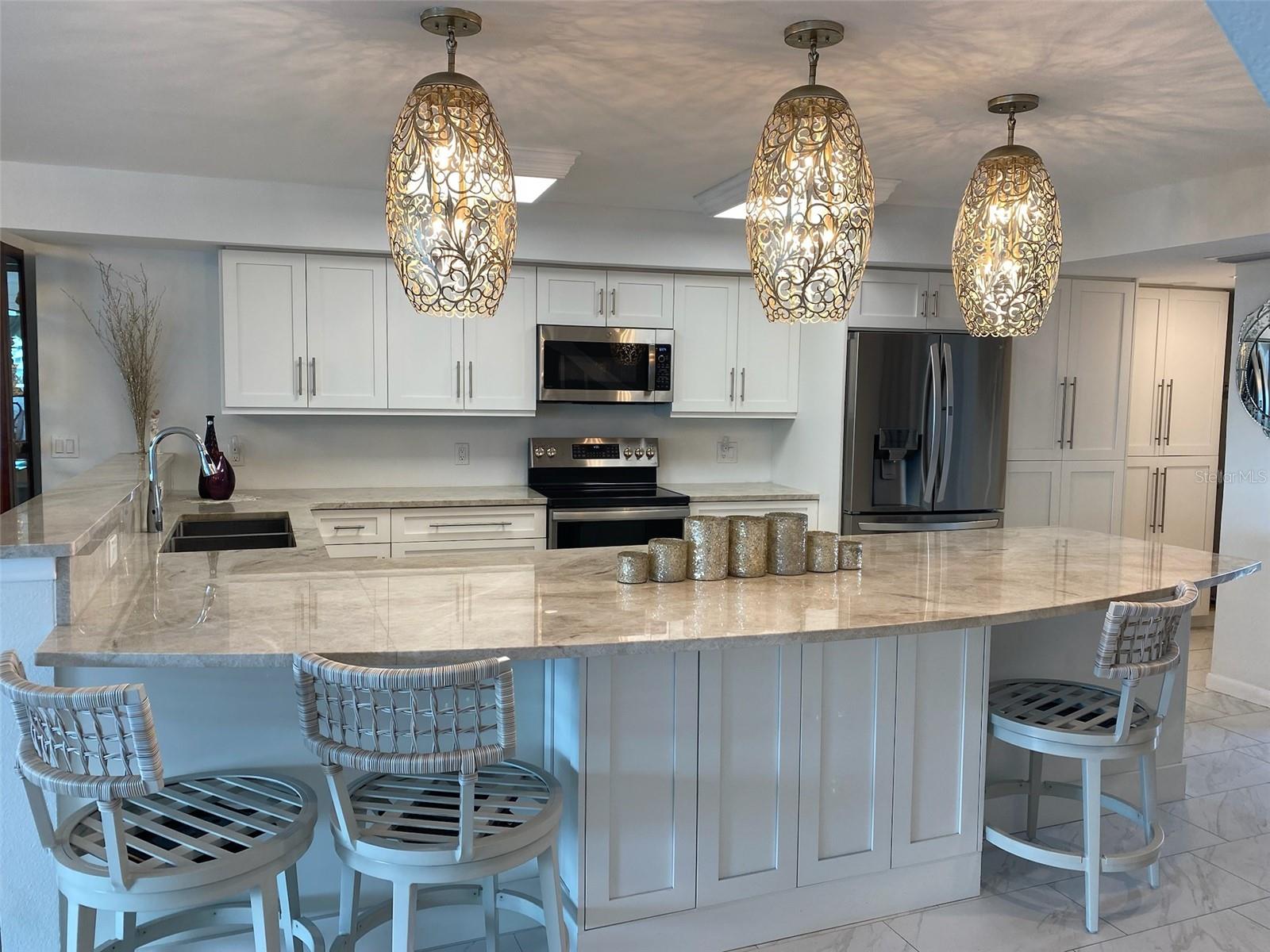 This beautifully updated kitchen features crisp white cabinetry, elegant marble countertops, and stainless steel appliances. Decorative pendant lighting adds a warm, inviting glow over the large breakfast bar, while modern seating provides the perfect spot for casual dining. The open layout and soft neutral tones create a bright, sophisticated space ideal for both cooking and entertaining.