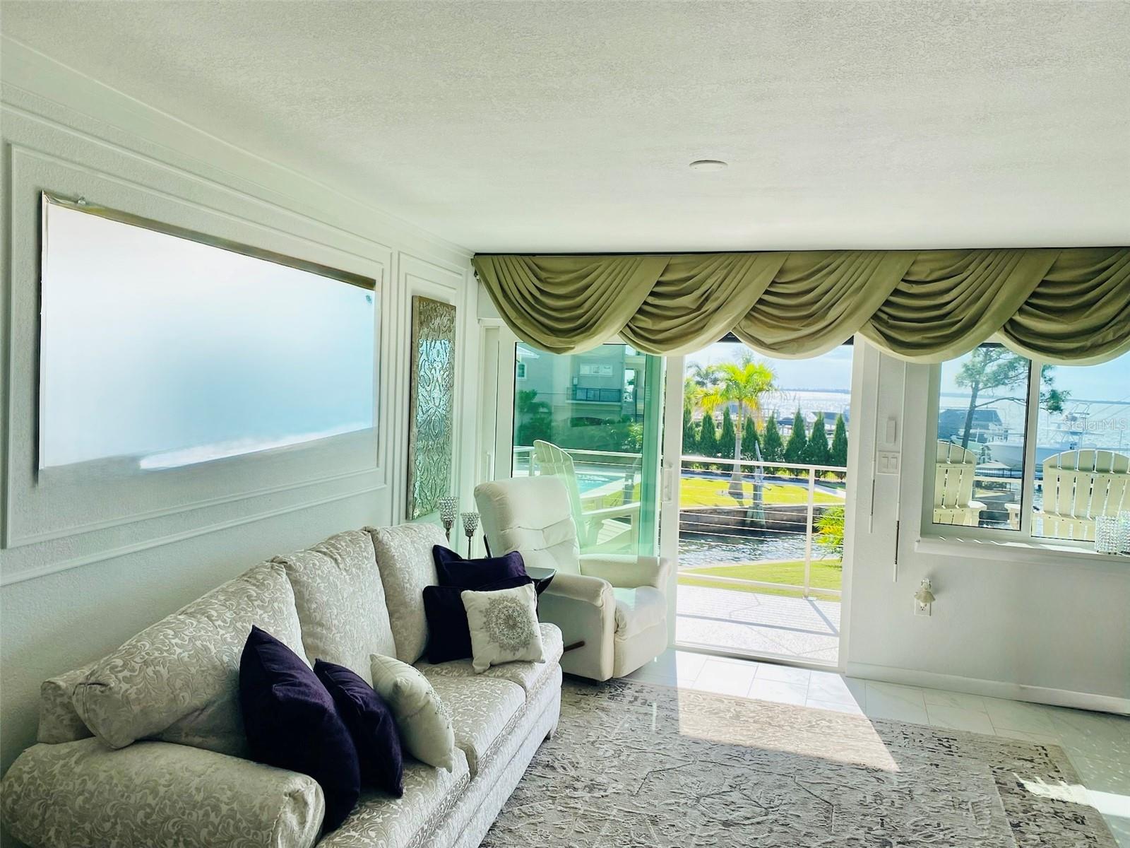 Bright waterfront living room featuring elegant drapery, cozy seating, and stunning views through expansive windows.