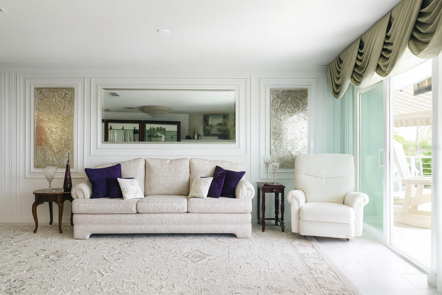 This bright and elegant living room features soft neutral tones, a beautifully patterned sofa, and plush seating that create a warm, inviting atmosphere. Decorative wall panels and a large framed mirror add depth and sophistication, while natural light floods the space through the sliding glass doors, highlighting the cozy yet refined design.
