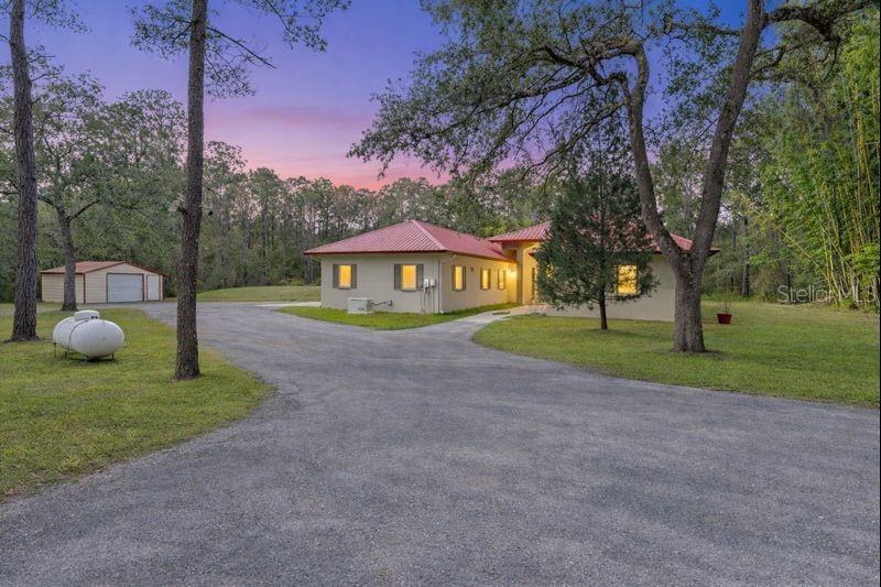At dusk, the home glows warmly against the surrounding trees, highlighting the peaceful, rural setting and expansive driveway.