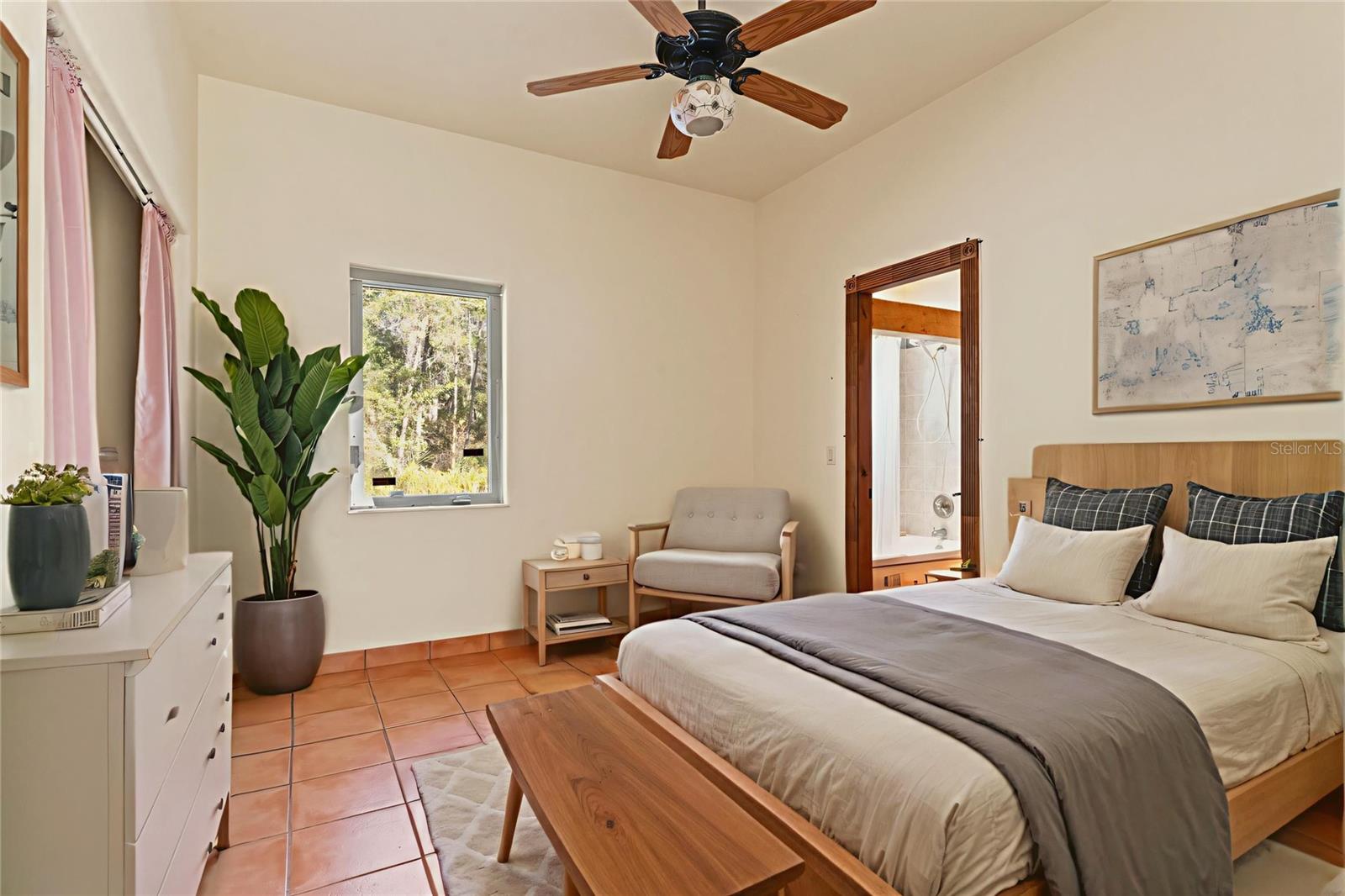 The serene guest room with Jack and Jill bath includes soft natural lighting, tile flooring, and a ceiling fan, with wooded views through large windows.