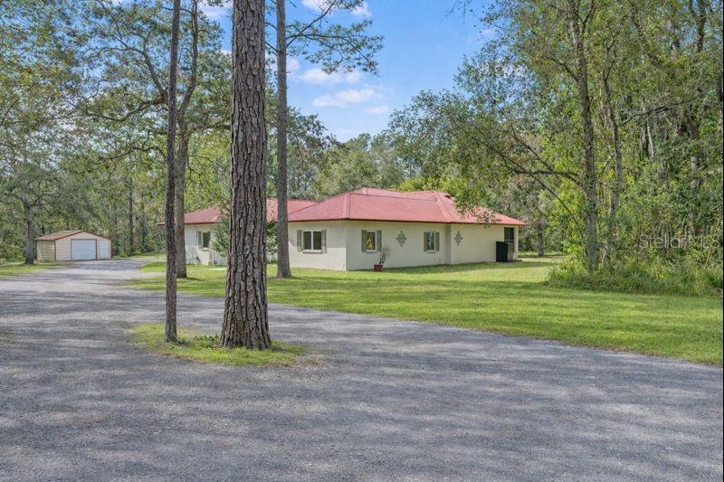 Nestled among mature trees, this charming single-story home features a red metal roof, cream-colored exterior, and detached garage, all set back from the road for added privacy.